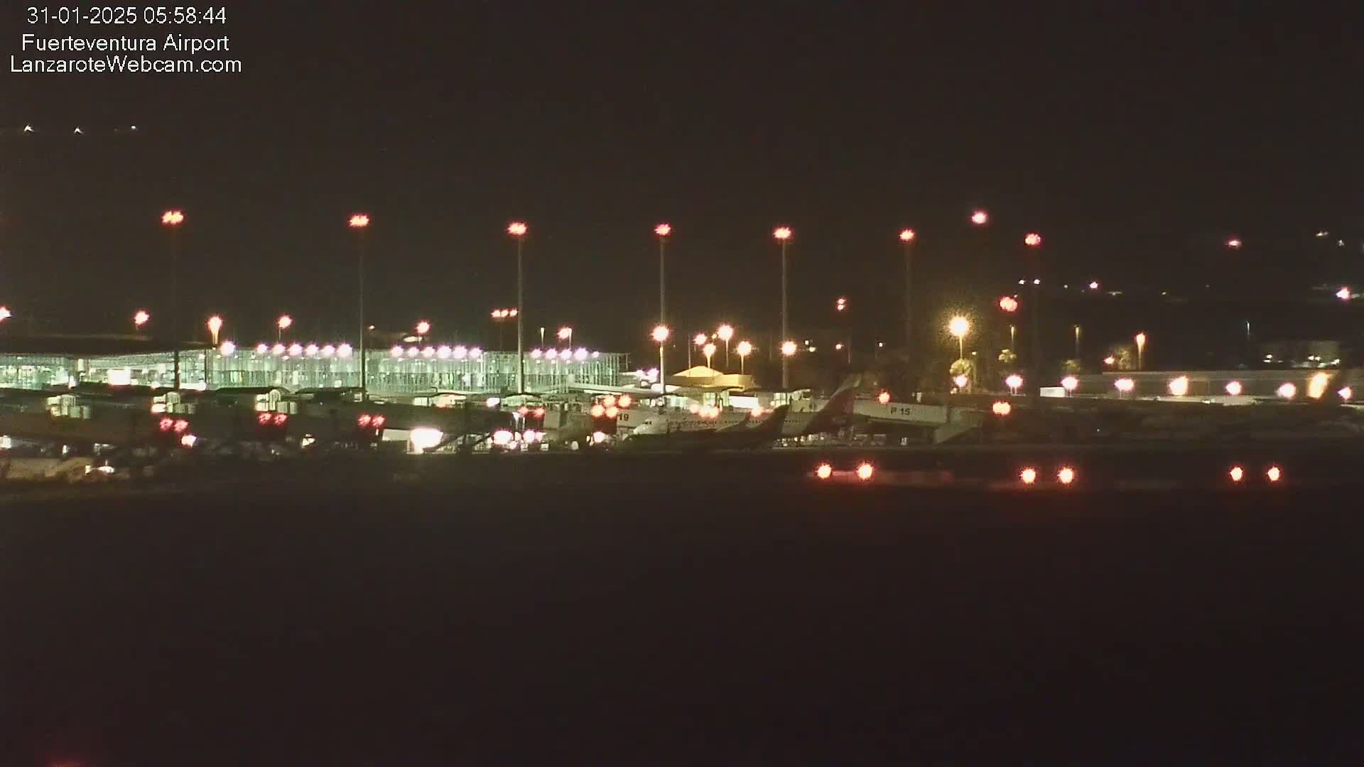 At night, several airplanes are parked on the tarmac near a brightly lit airport terminal under a clear, dark sky.