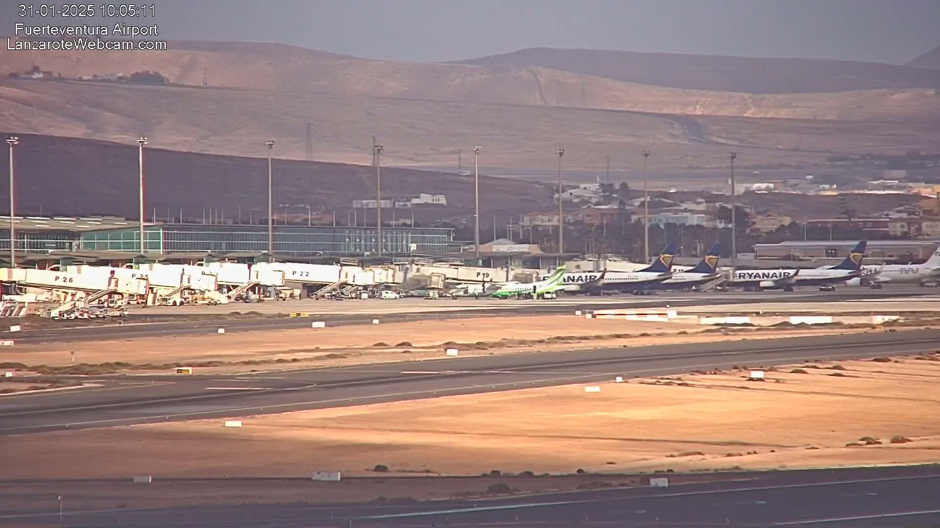 Several airplanes are parked at an airport tarmac under a hazy sky, with a mountainous landscape visible in the background.