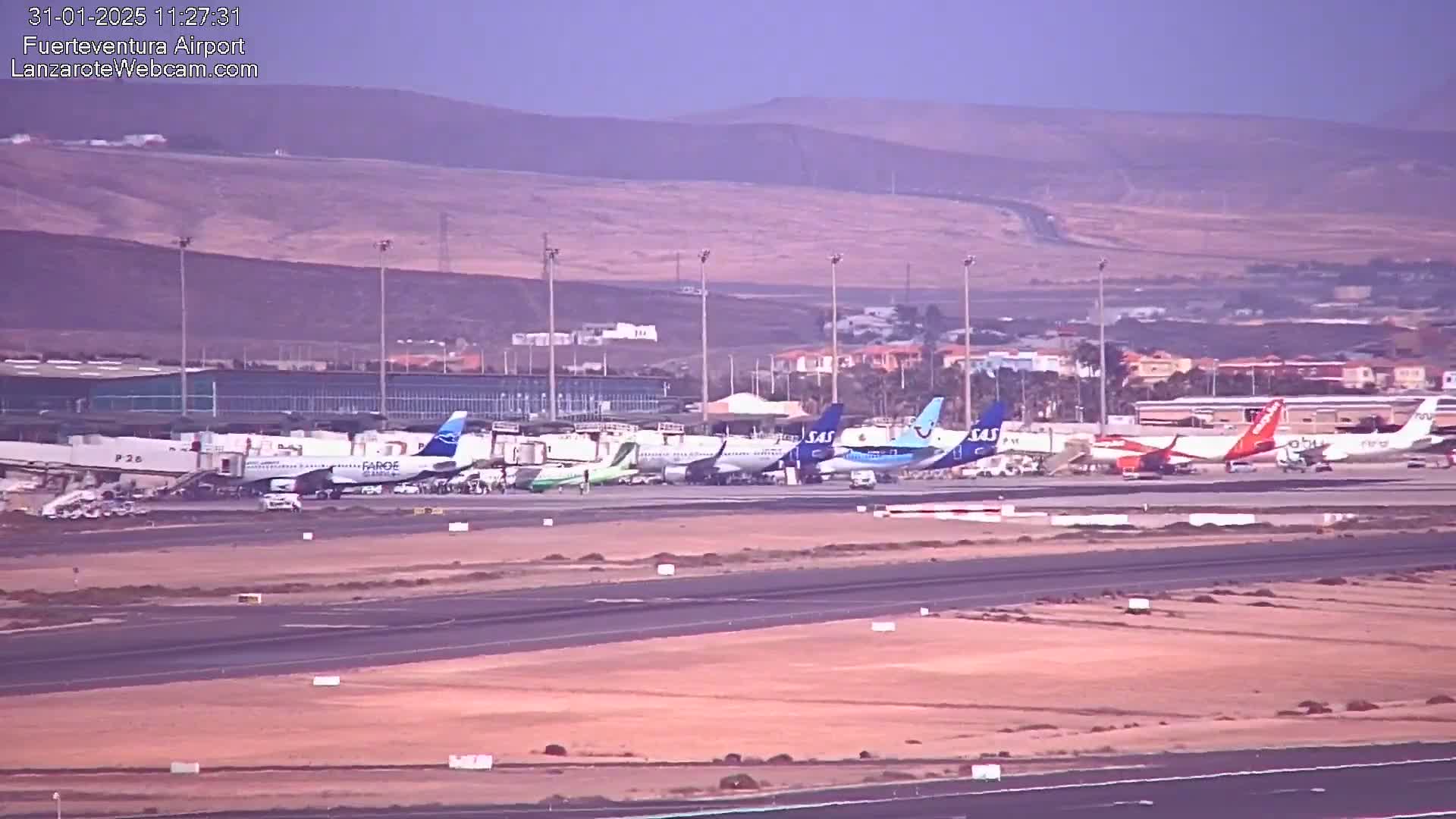 Several airplanes are parked at an airport tarmac on a hazy day with mountains visible in the background.