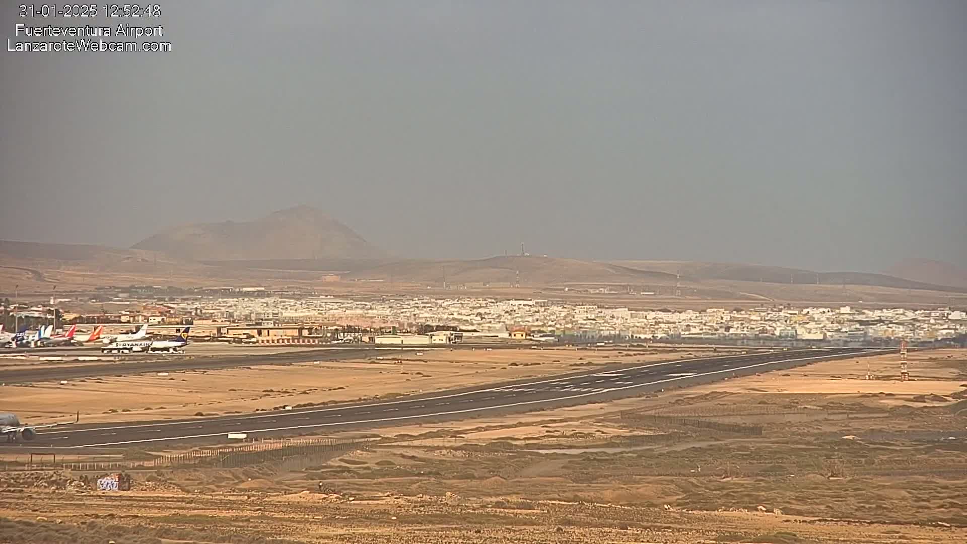 A long runway at an airport is visible, with several airplanes parked nearby, a town in the distance, and hazy weather conditions.