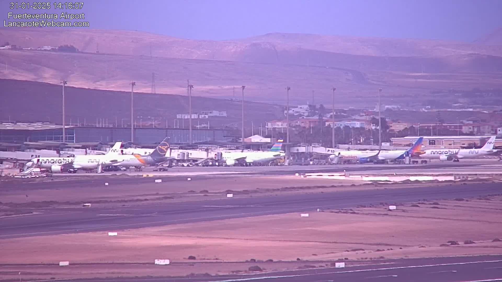 Several airplanes are parked at a terminal at an airport with hazy, muted-colored mountains in the background.