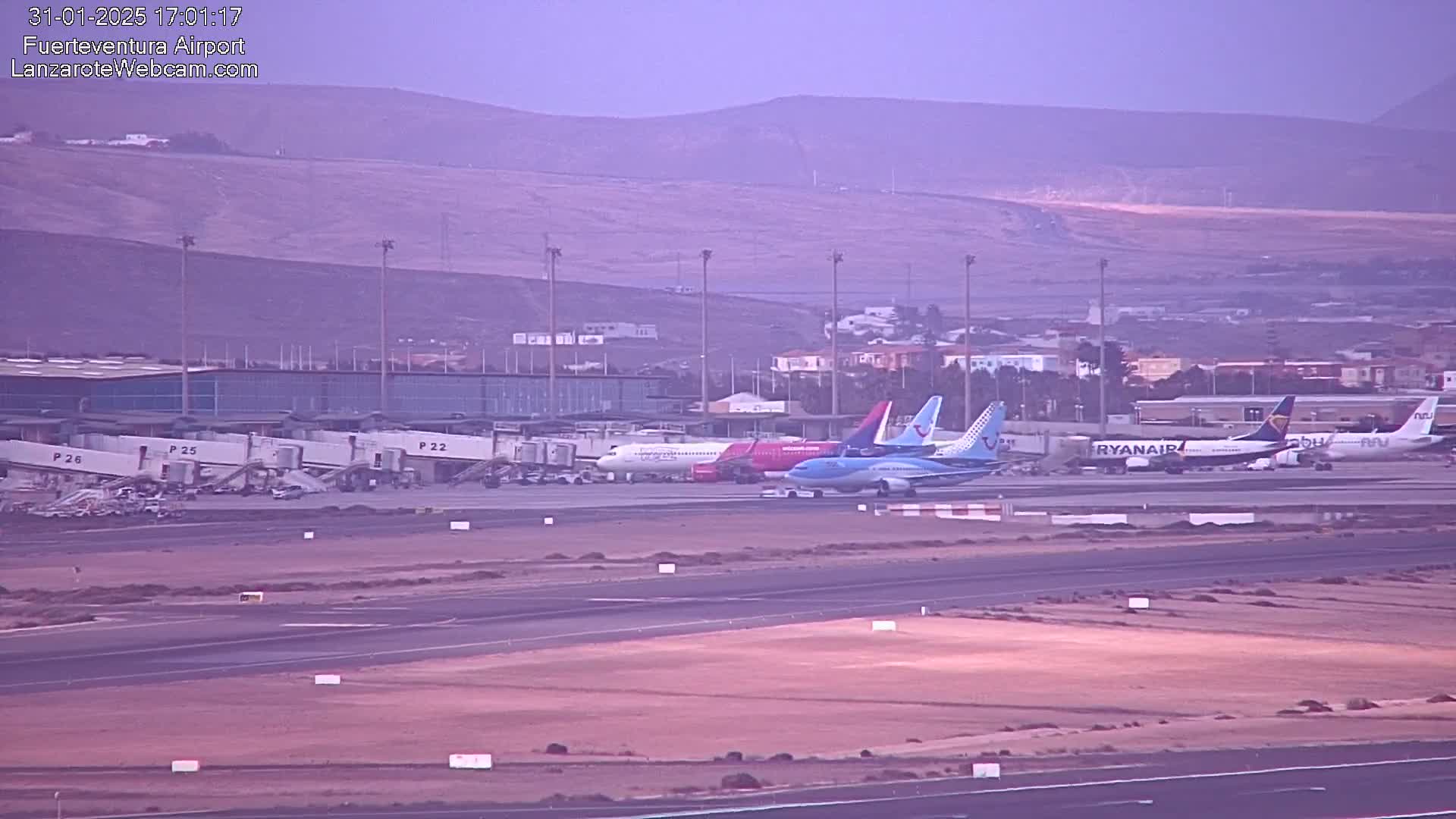 Several airplanes are parked at an airport tarmac under a hazy, light-purple sky, with a distant mountain range visible in the background.