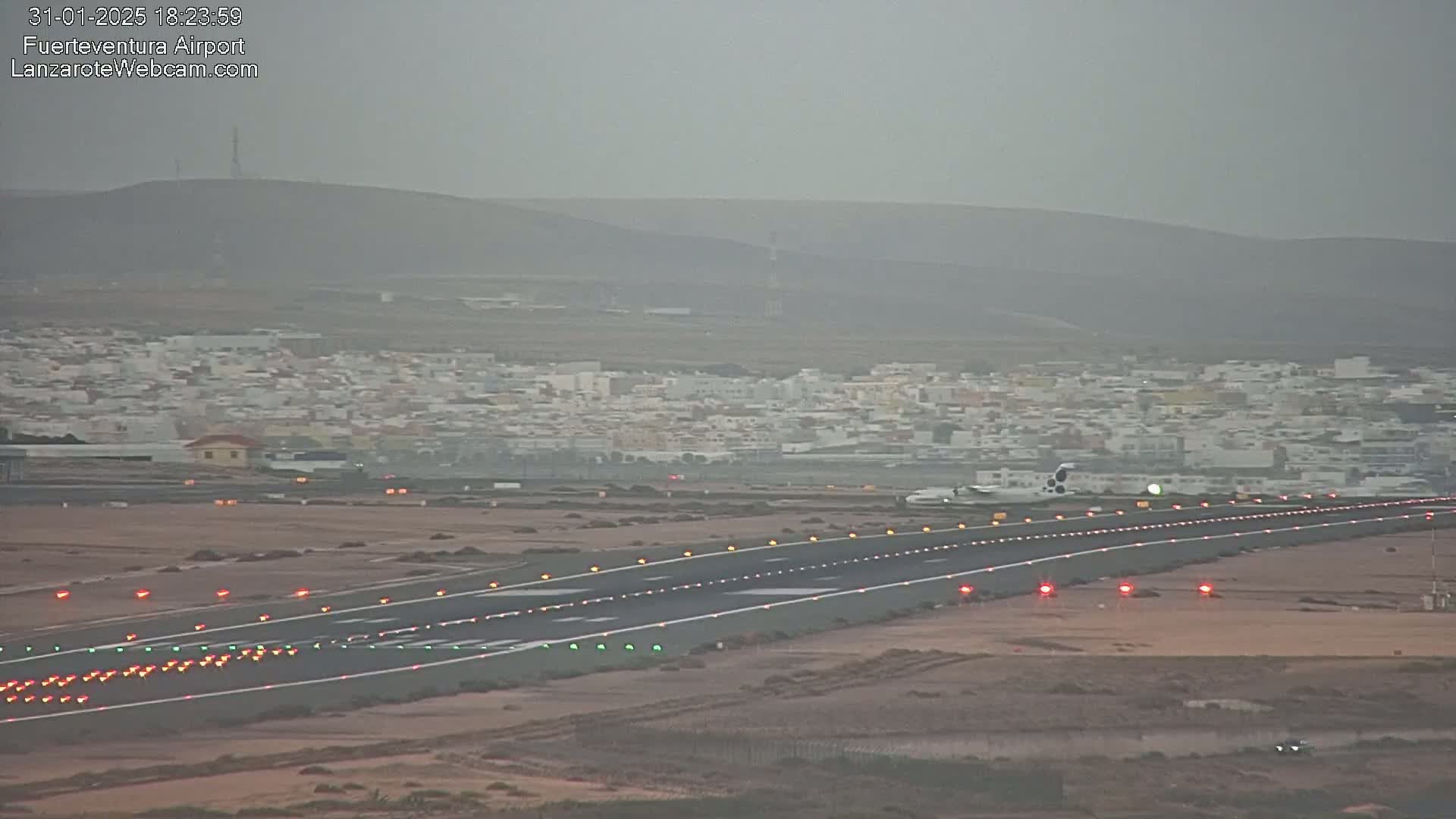 A hazy view shows an airplane taxiing on a runway towards a town nestled at the base of a distant hill.