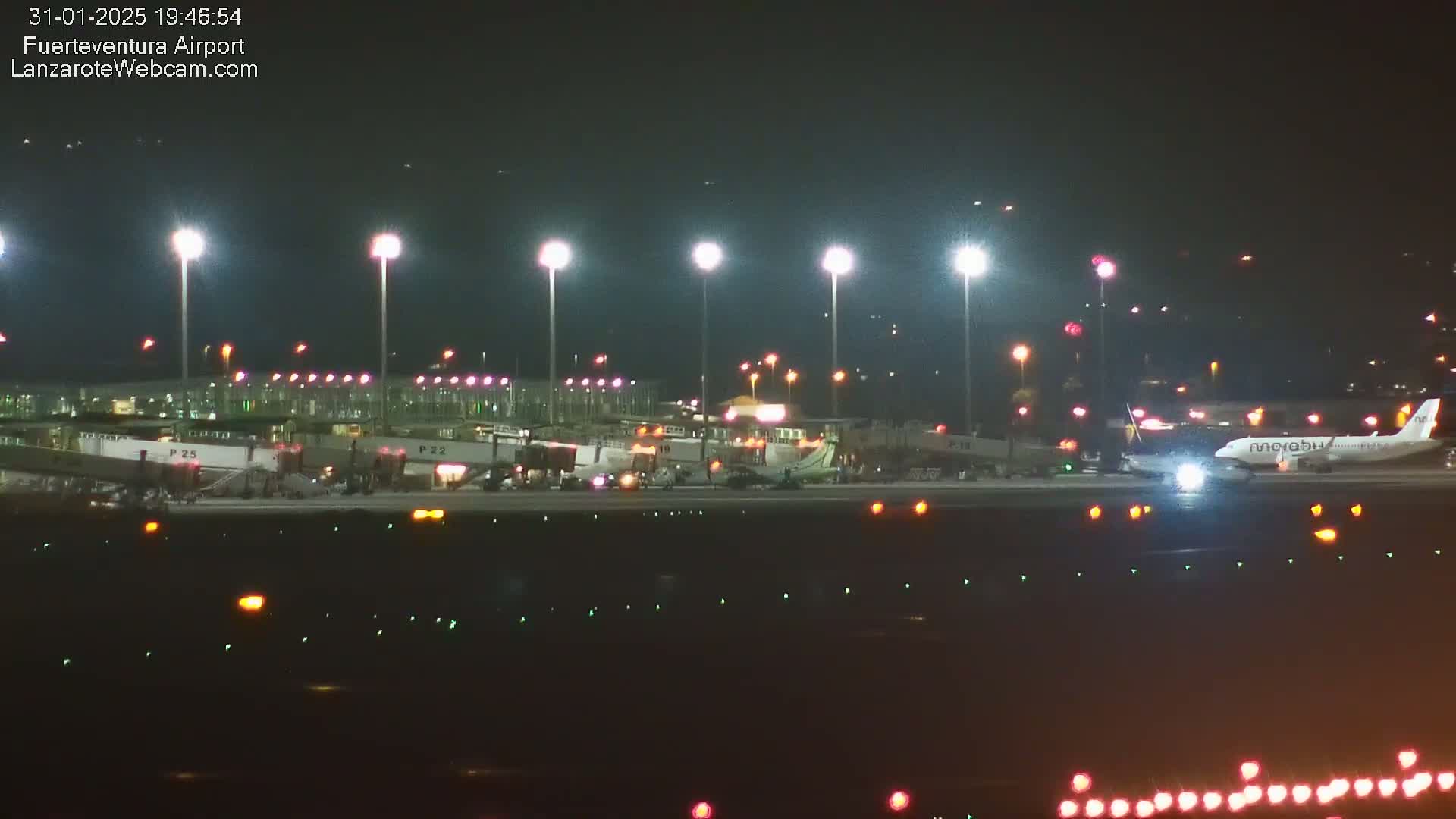 A brightly lit airport tarmac at night shows several planes near gates and one plane on the runway.
