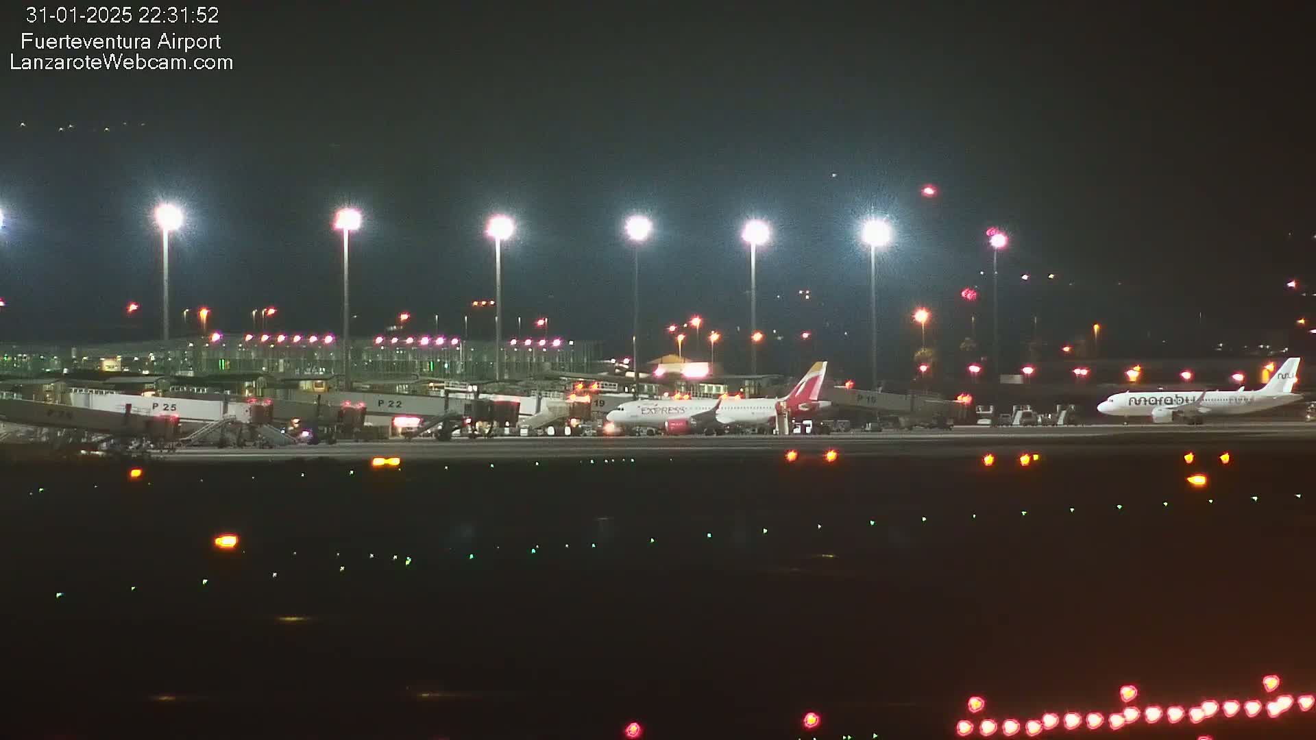 At night, two airplanes are parked at gates at an airport with bright lights illuminating the tarmac.