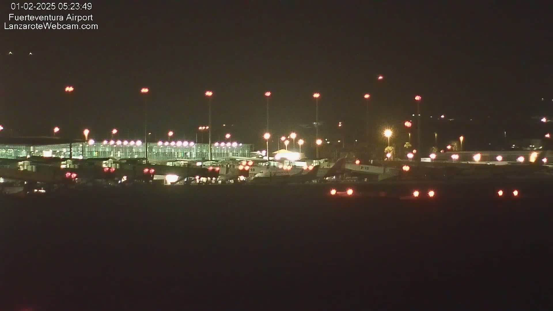 A nighttime view of an airport tarmac with several airplanes parked near a brightly lit terminal building under a clear, dark sky.
