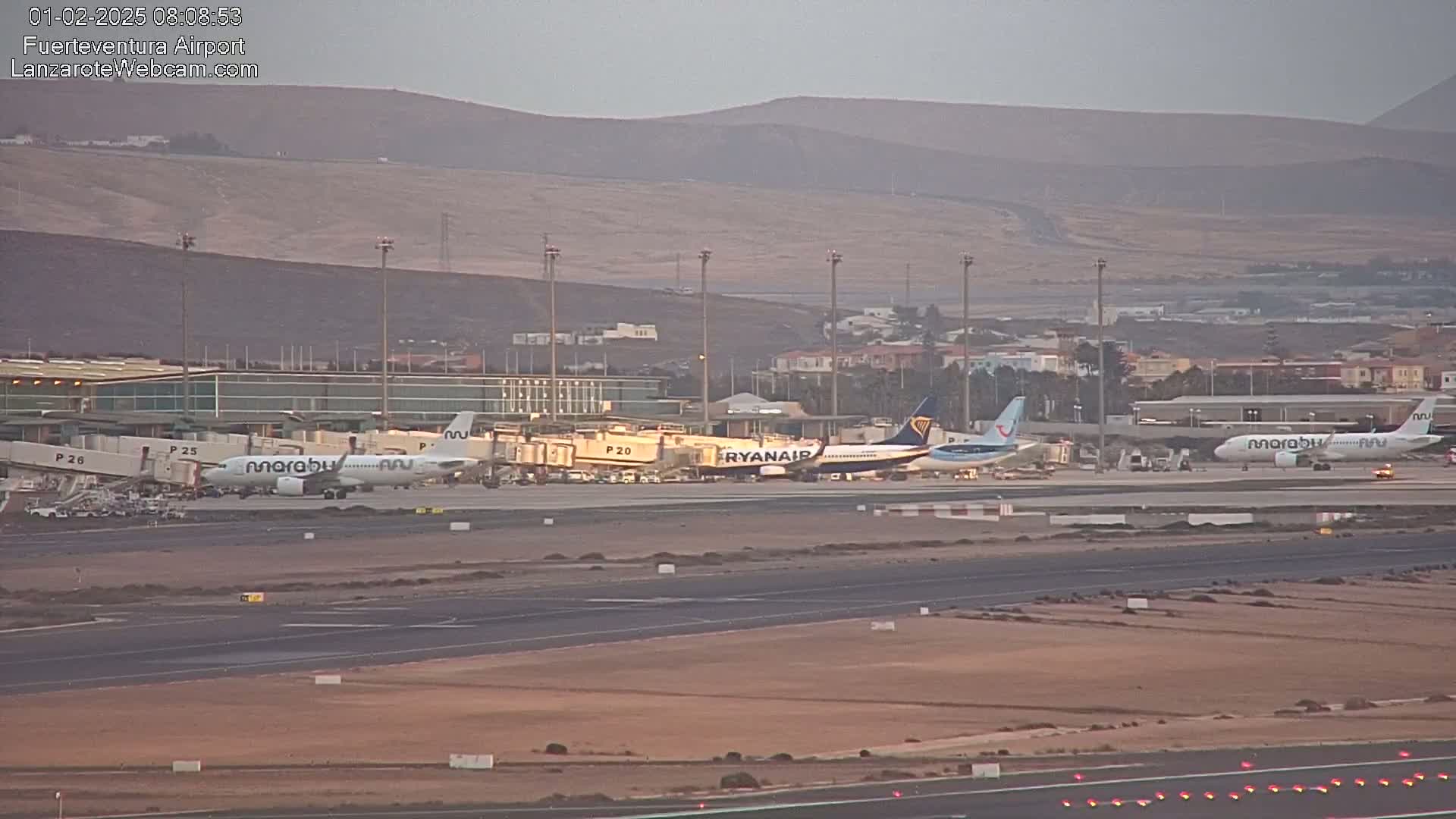 Several airplanes are parked at an airport tarmac under a hazy sky with distant, low-lying hills in the background.