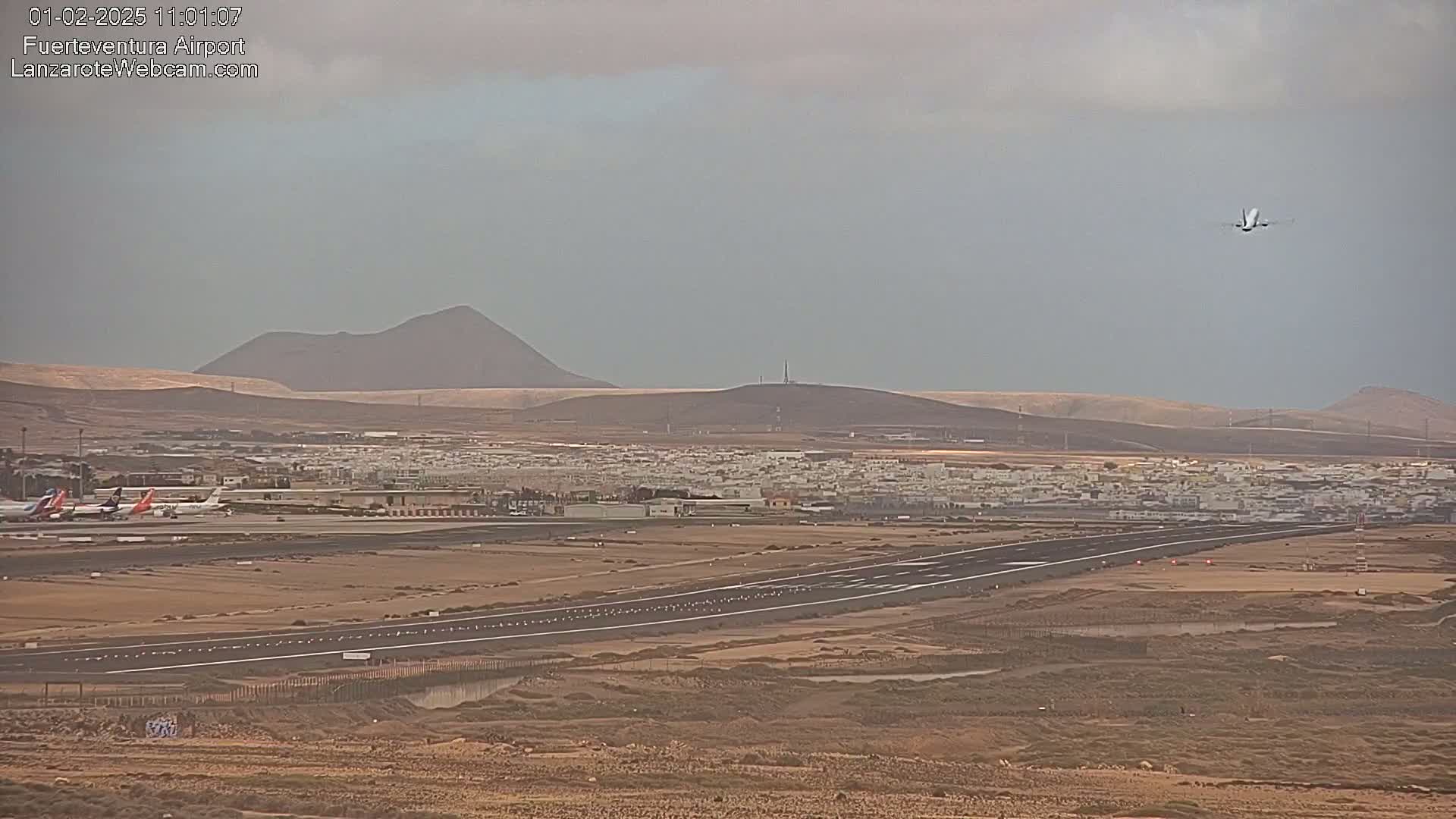 An airport runway with several planes on the tarmac and one plane taking off is visible beneath a gray, overcast sky, with arid, mountainous terrain in the background.