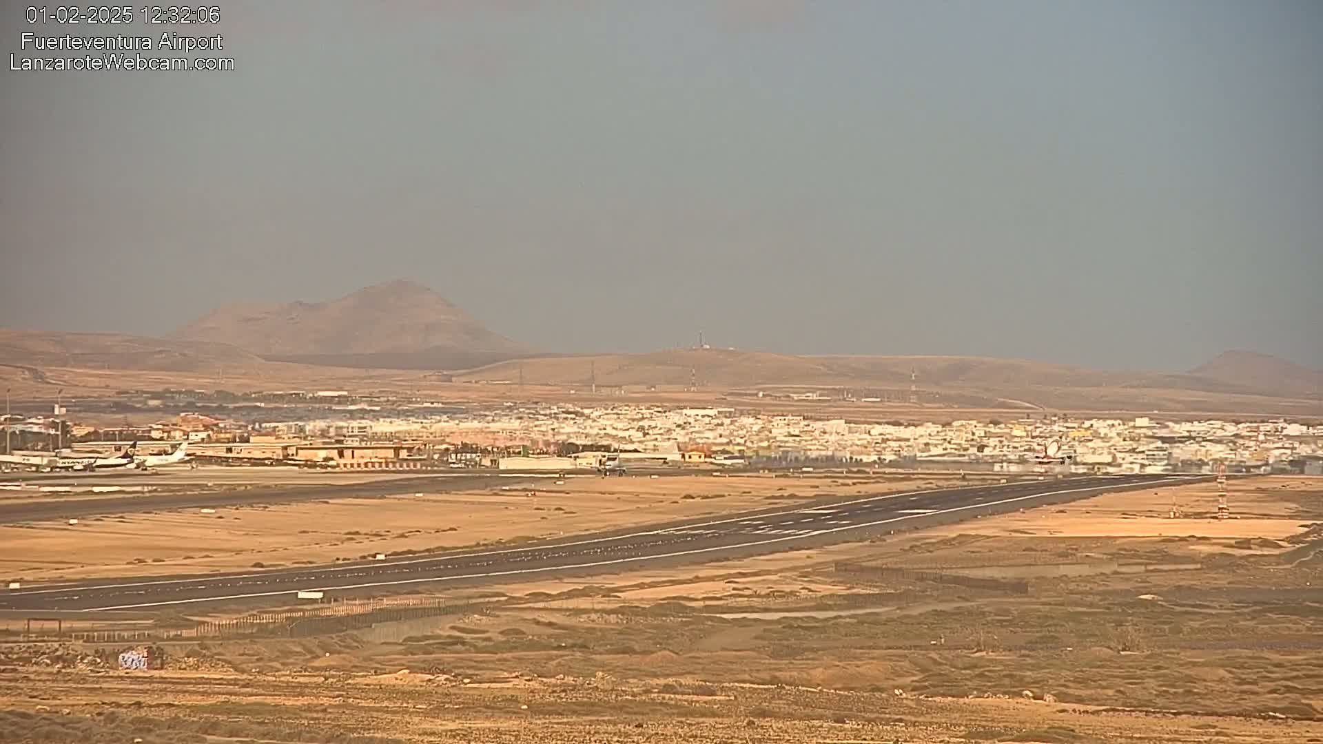 A hazy, sunny day reveals an airport runway, with a plane on the tarmac and a town in the background, all situated before a line of low, arid hills.