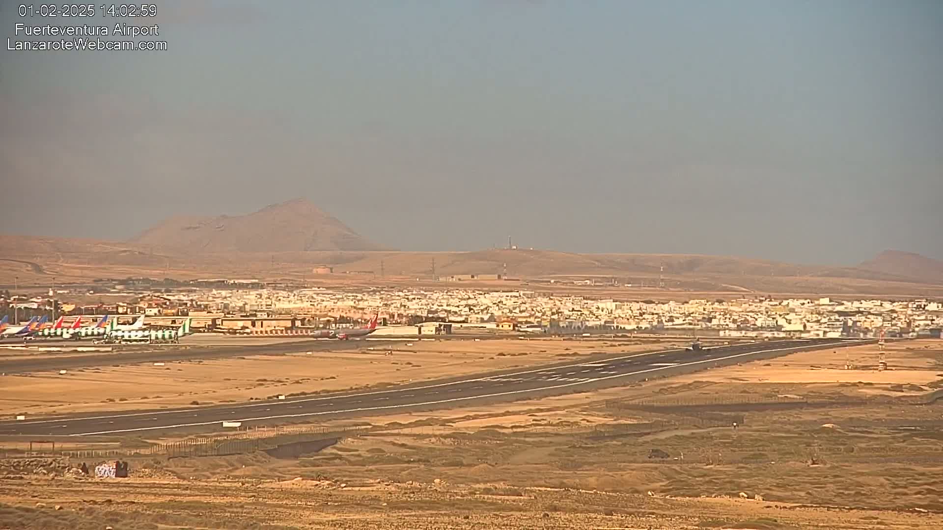 A long runway at an airport is visible, with several airplanes parked near a town nestled against hazy brown hills under a clear, light-grey sky.