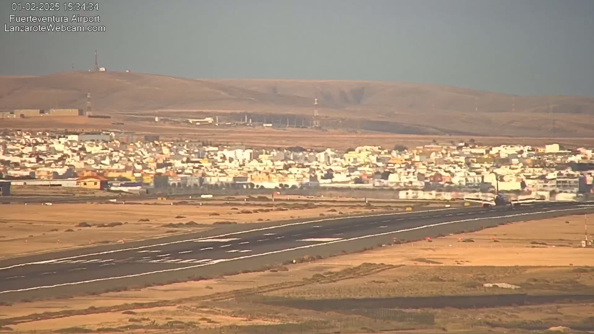 A plane is landing on a runway near a town under hazy, sunny conditions, with hills visible in the background.