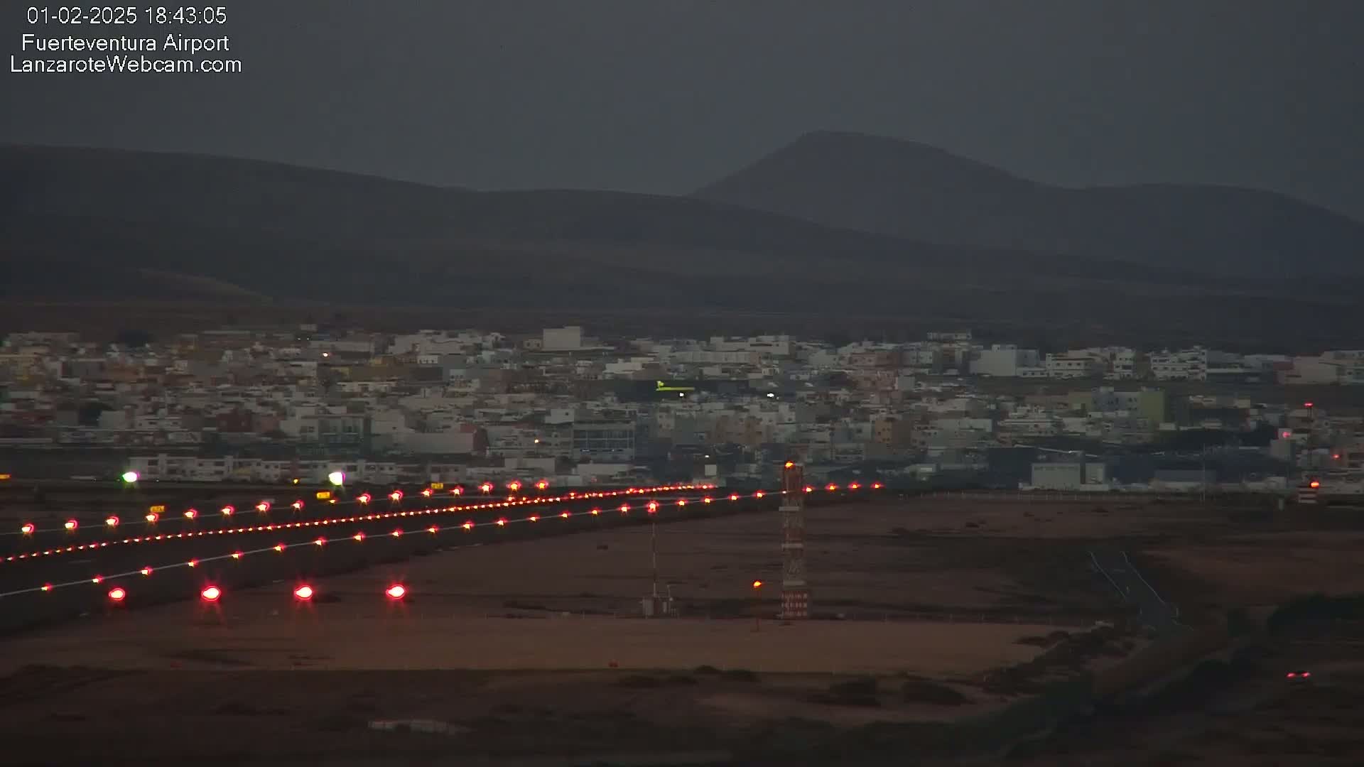 A dark, twilight-lit airport runway with red lights illuminates a town nestled against dark hills in the background.