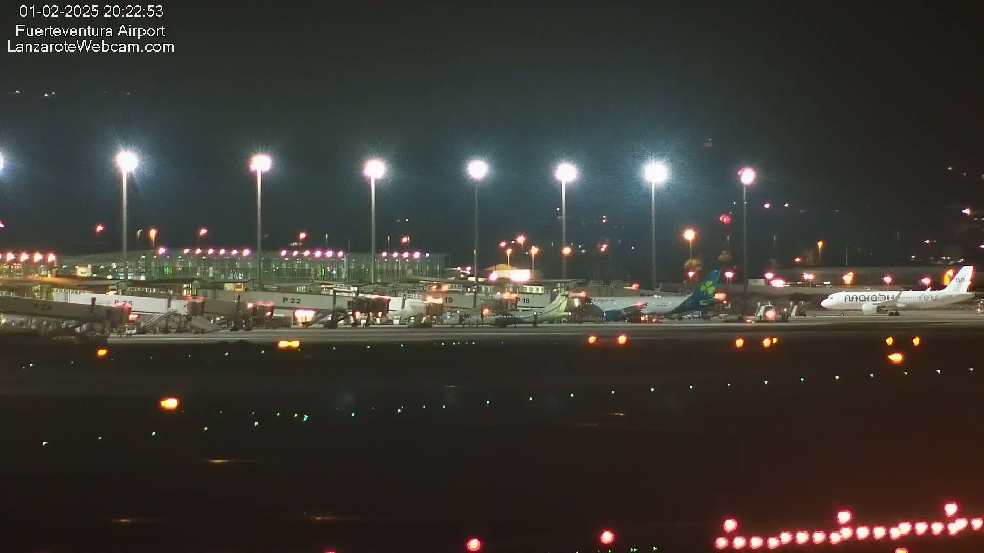 At night, brightly lit airplanes are parked at gates at an airport with jet bridges attached.