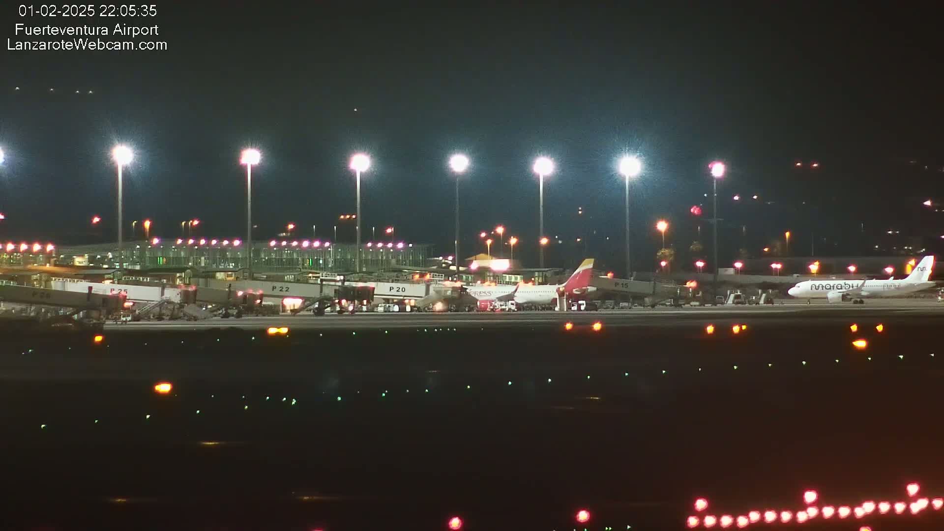 At night, several airplanes are parked at gates at an airport, brightly lit by floodlights.