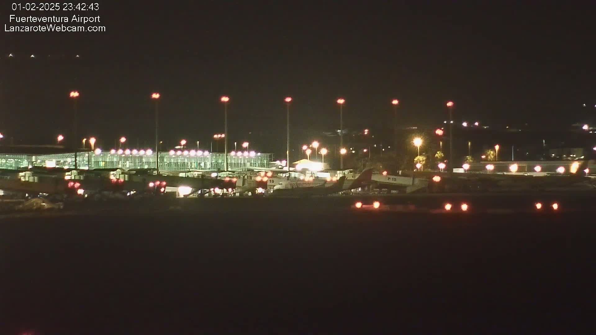 A nighttime view of an airport terminal and tarmac, illuminated by numerous lights, with several airplanes visible near the gates.