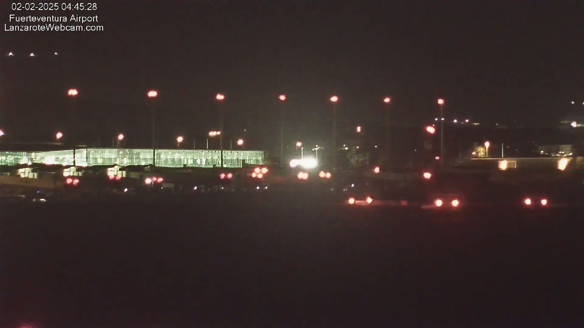 At night, a long, low building with lights is visible beyond a roadway with several vehicles whose taillights are illuminated.