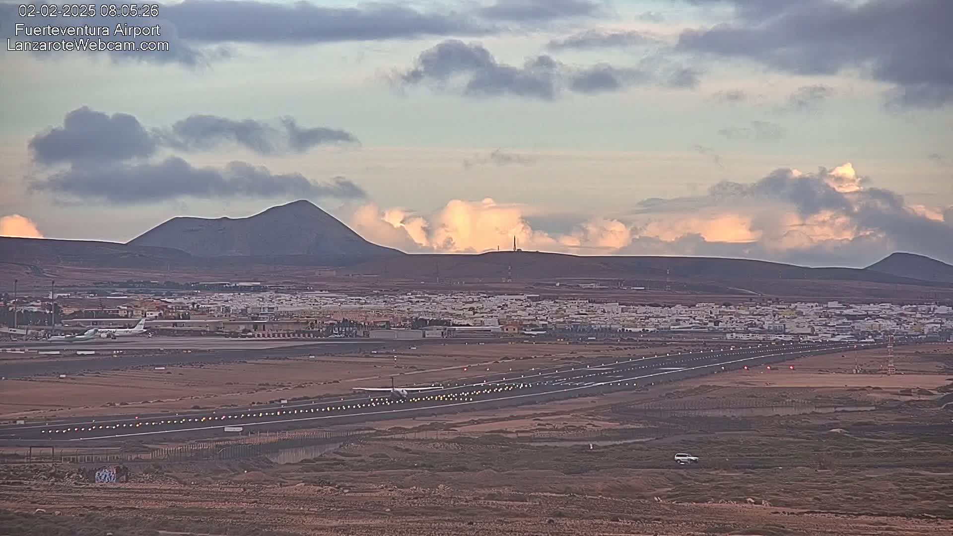 A small airplane taxis on a runway at an airport near a town nestled amongst volcanic hills under a partly cloudy sky.