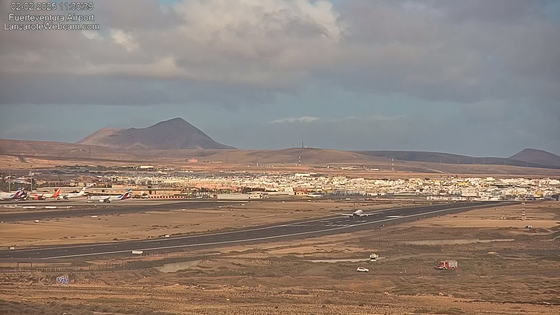 A plane taxis on a runway at an airport near a town, with several other planes parked nearby, under a partly cloudy sky and arid, mountainous landscape.