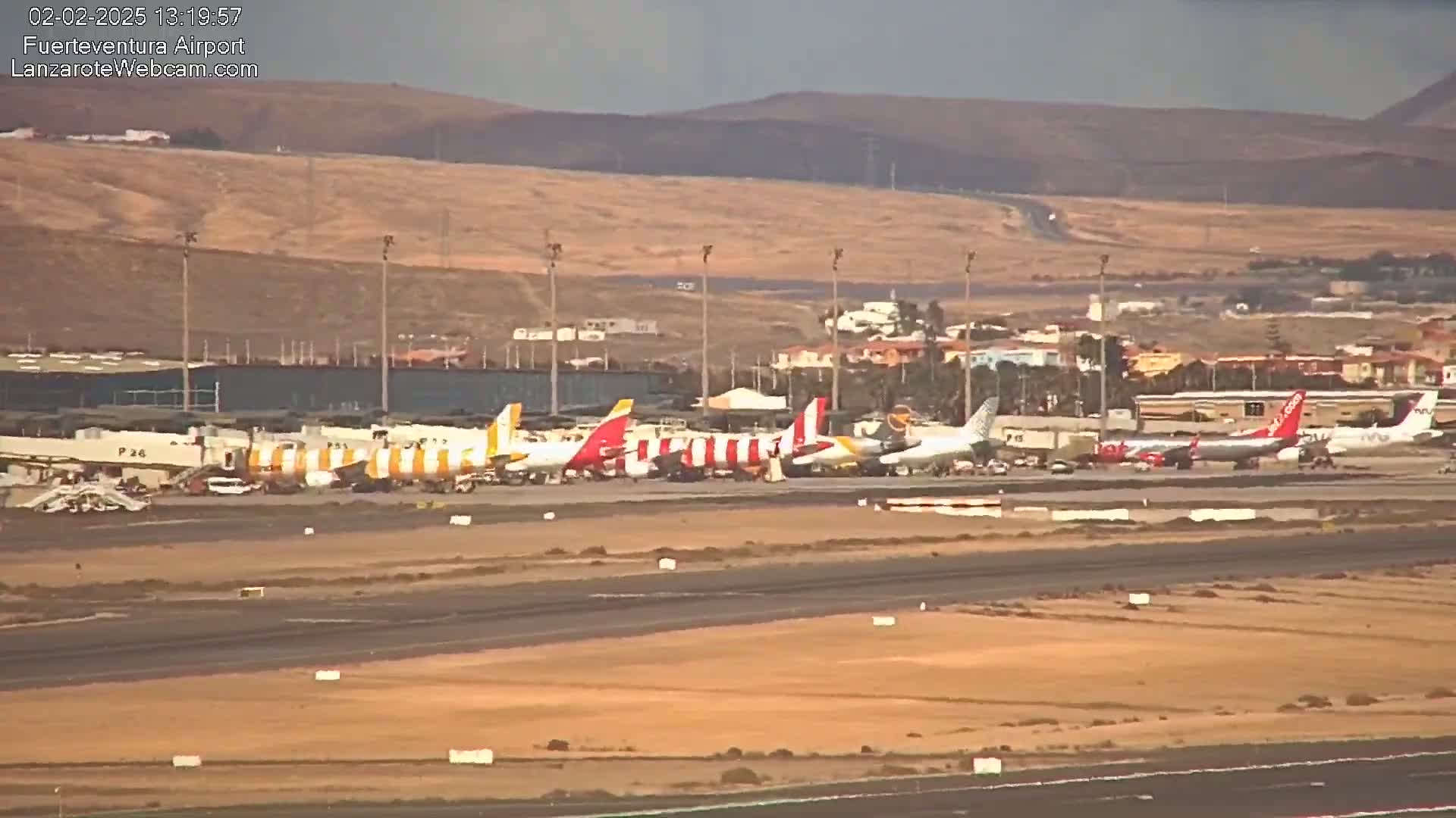 Several airplanes are parked on an airport tarmac under a hazy sky, with a hilly, desert-like landscape in the background.