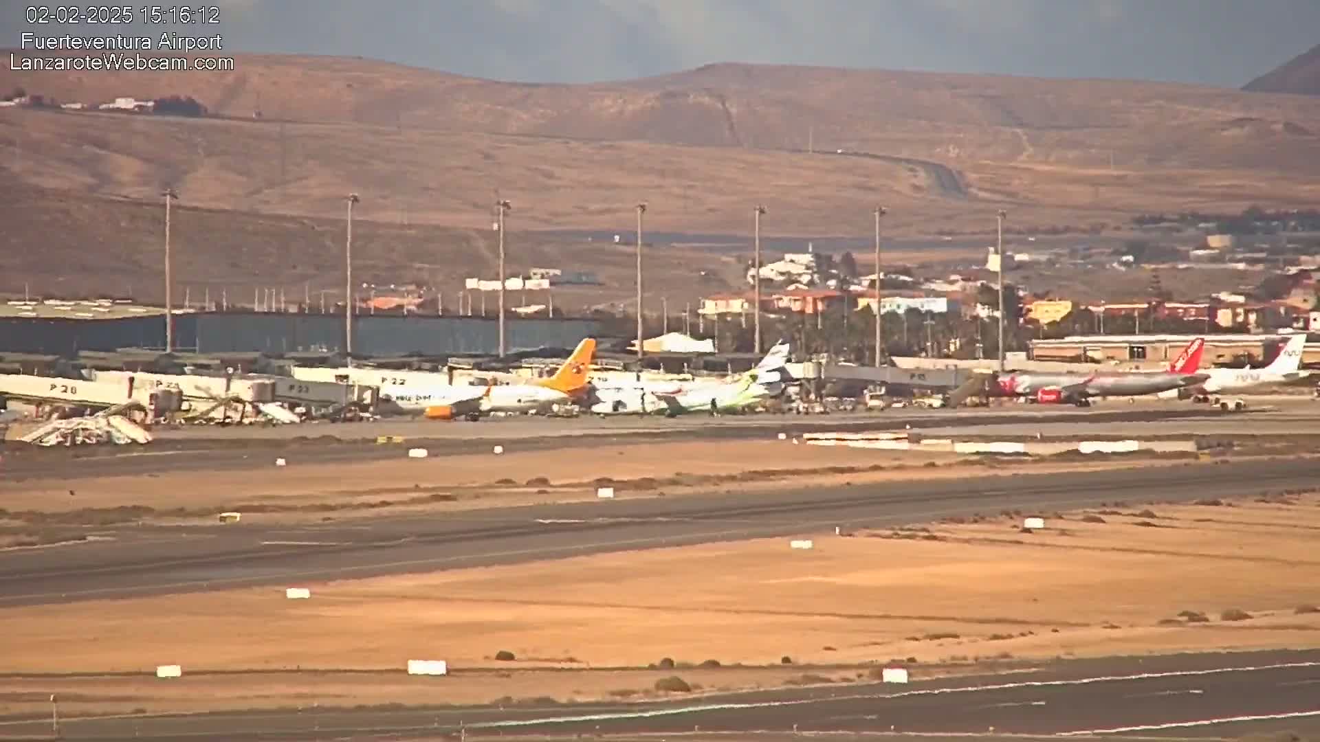 Several airplanes are parked at an airport tarmac under partly cloudy skies, with a distant view of brown hills.