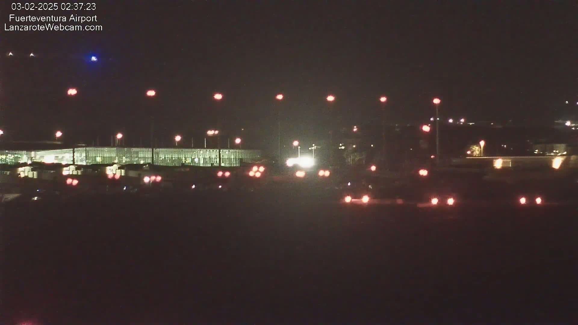A nighttime view of an airport tarmac with several lights illuminating vehicles and the terminal building under a clear, dark sky.