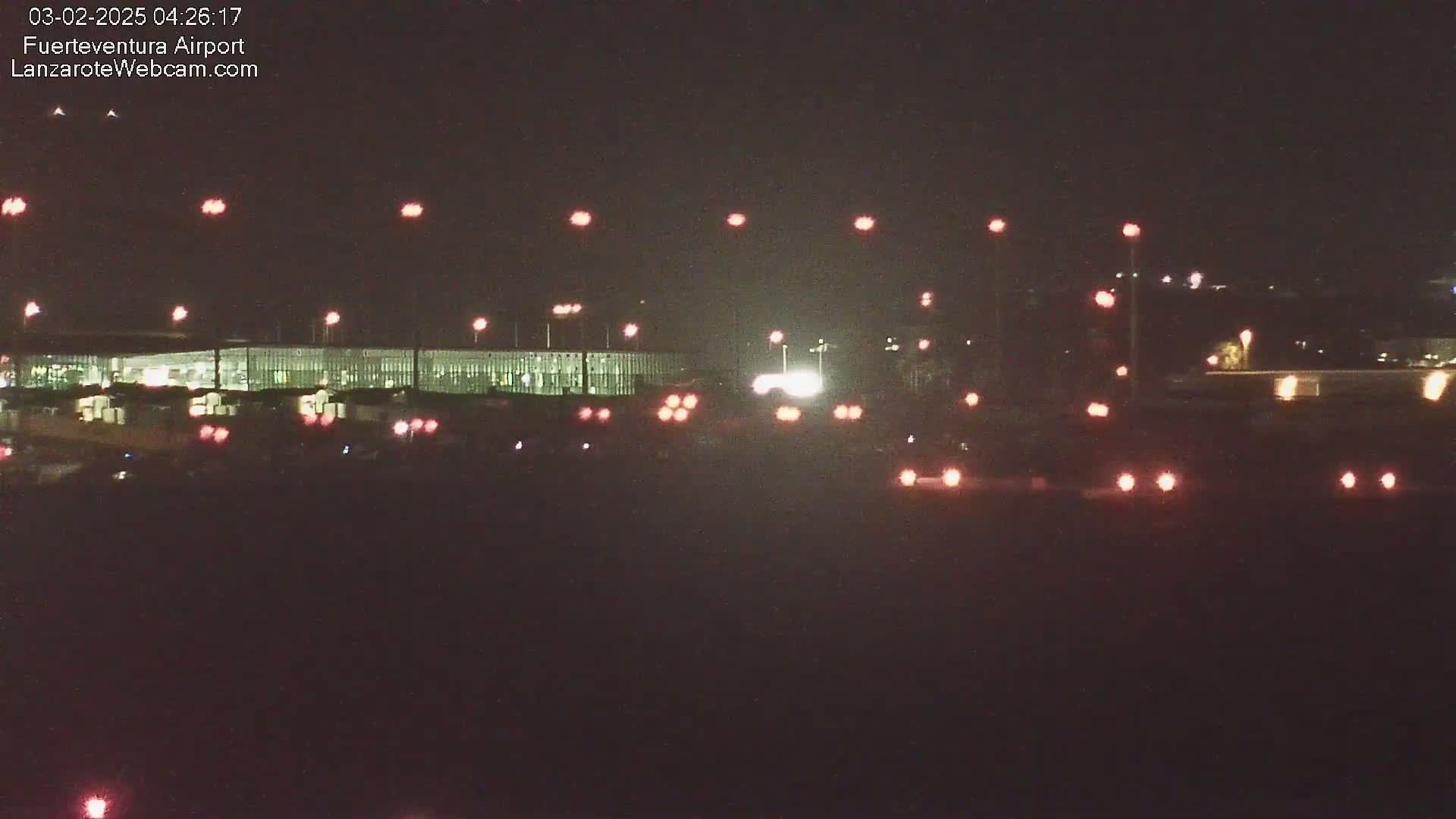 At night, numerous red and white lights illuminate a long, low building, likely an airport terminal, with various vehicles visible in the foreground.