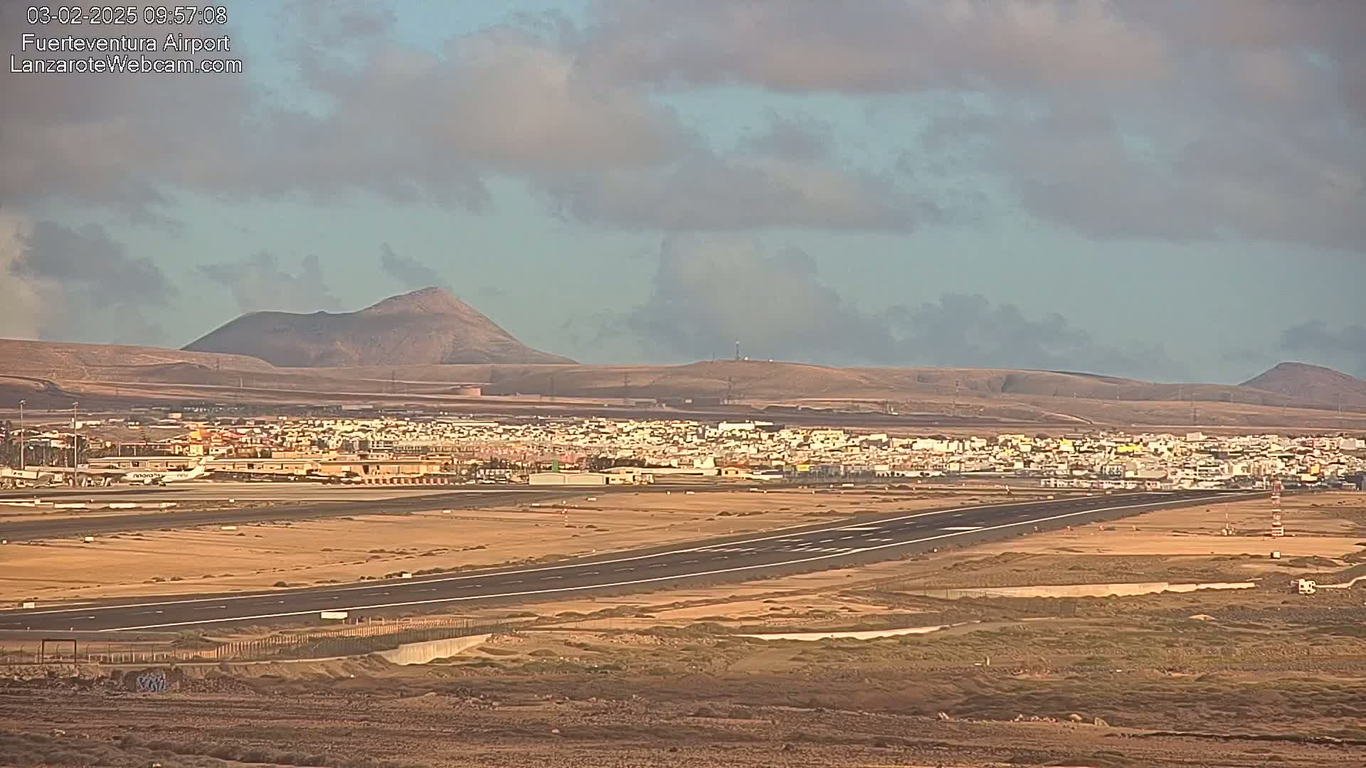 A runway at an airport, situated near a town and backed by volcanic hills under a partly cloudy sky.