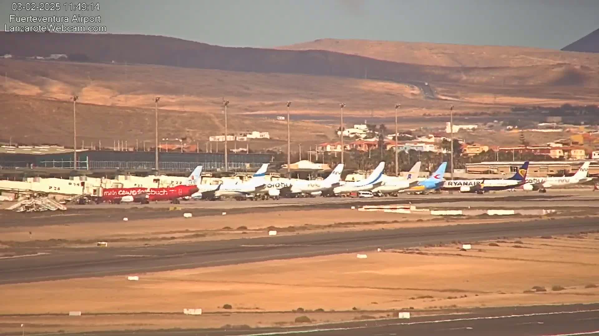 Several airplanes are parked at an airport tarmac under a partly cloudy sky, with arid hills visible in the background.