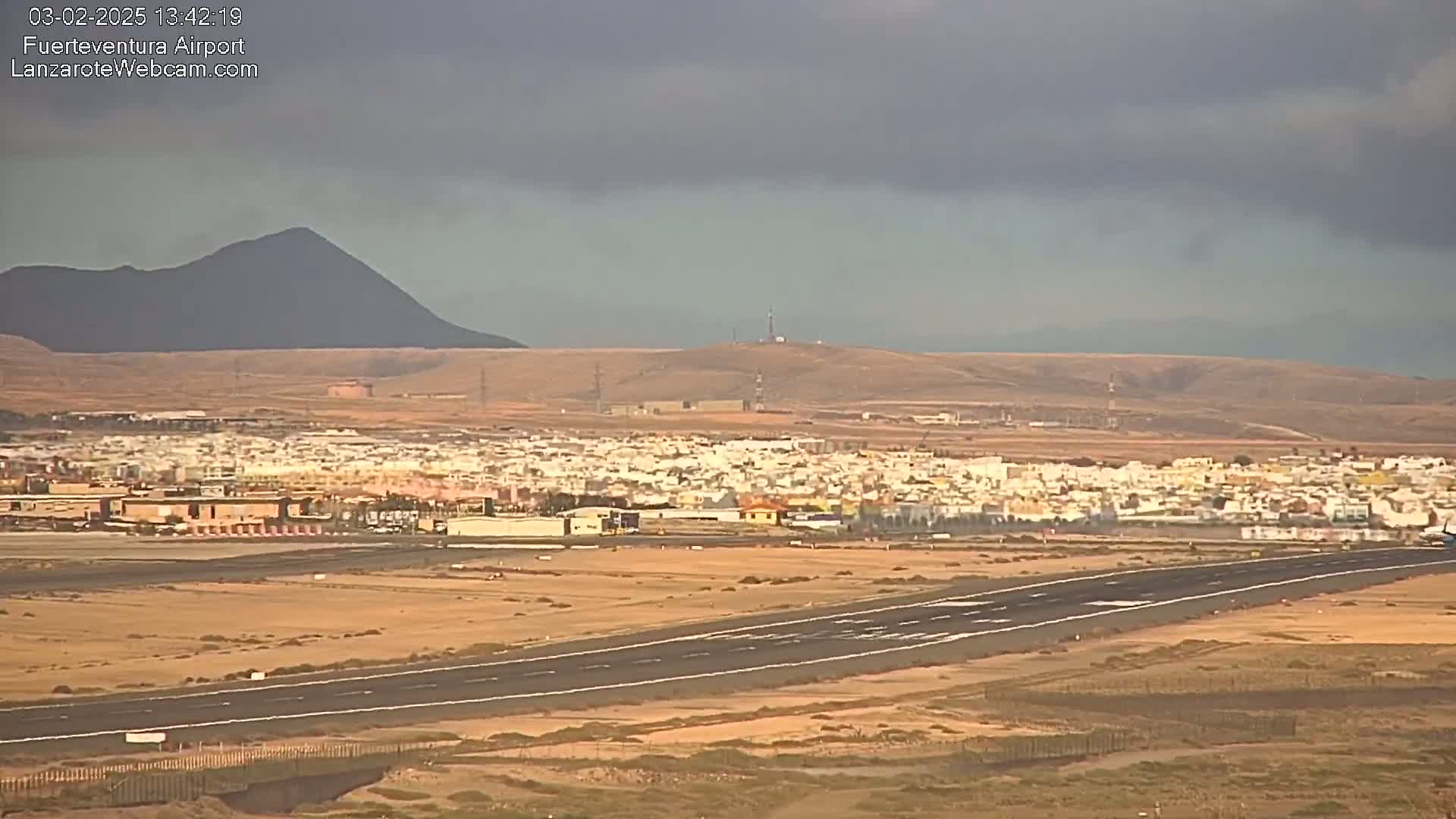 An airport runway stretches across a flat, arid landscape under a cloudy sky, with a town and distant mountains in the background.