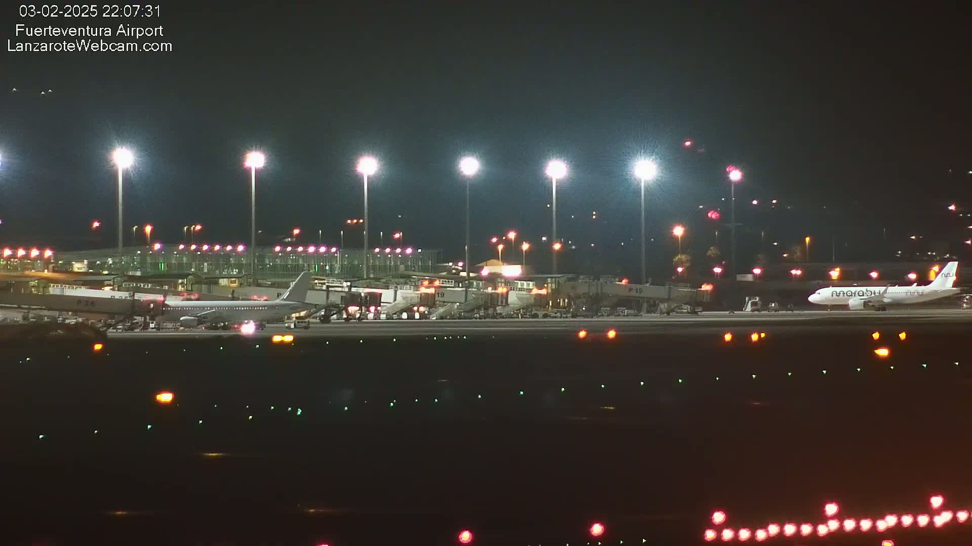 A nighttime view of an airport tarmac, showing an airplane parked near a terminal building under bright lights.
