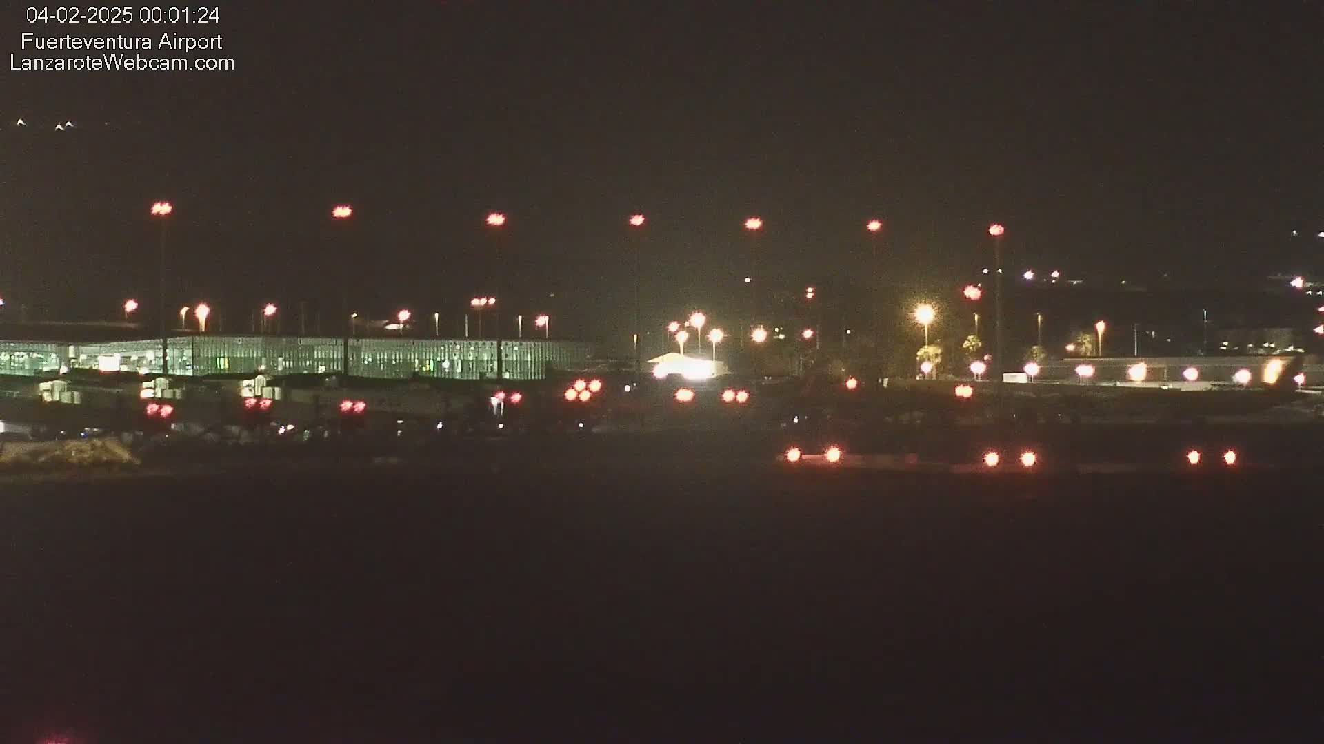 A nighttime view of an airport terminal building illuminated by numerous lights, with vehicles visible in the foreground.