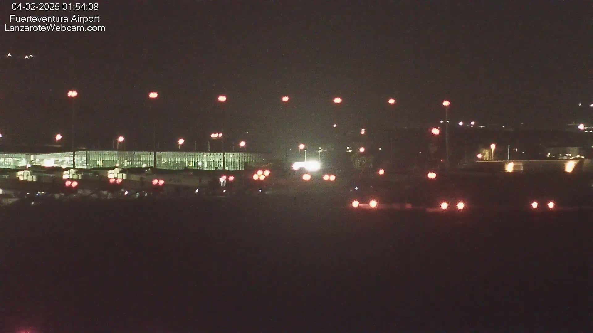 At night, a long, low building is illuminated, with various lights scattered across a dark area in front of it.