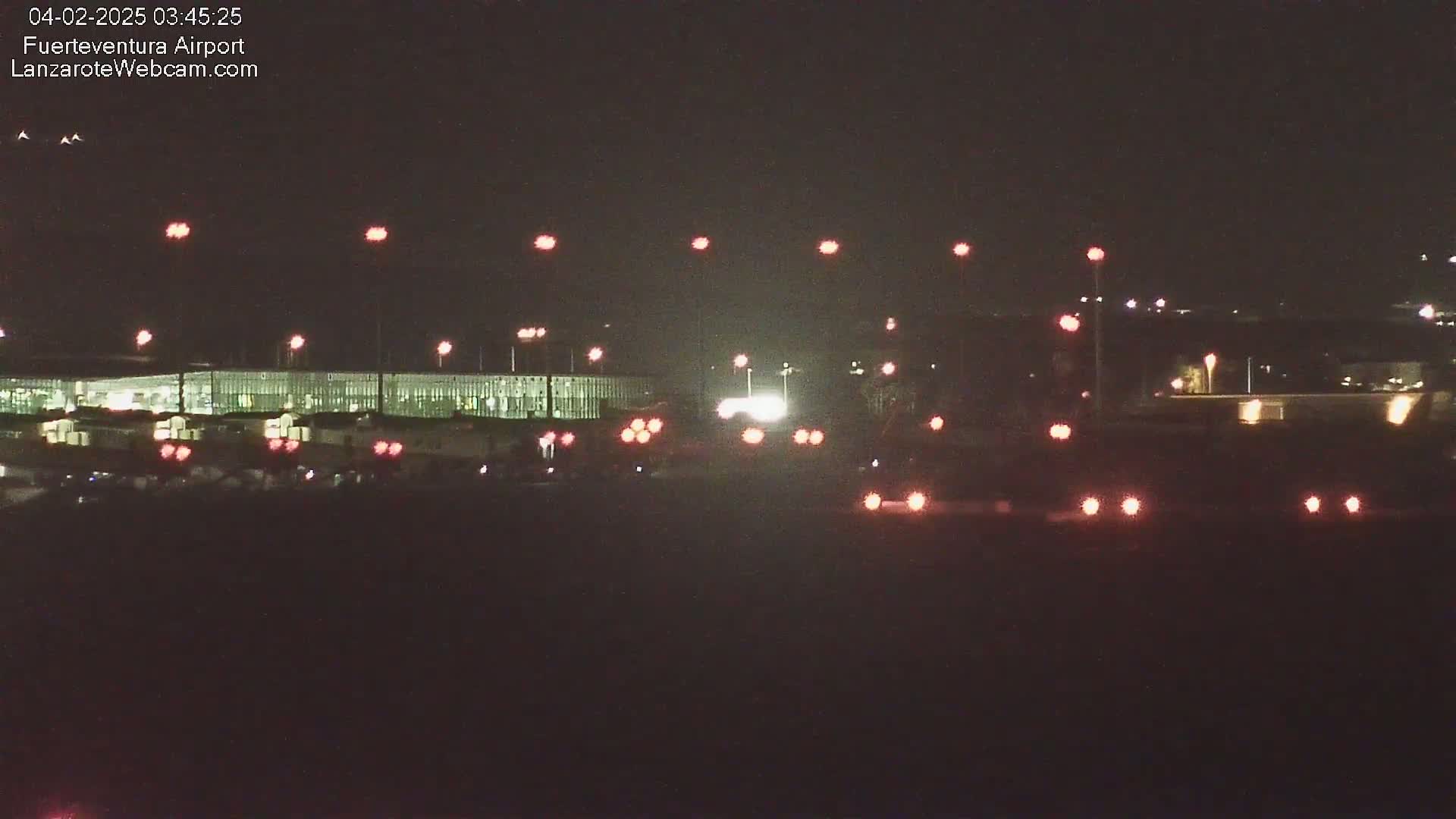 At night, a long building with lights is visible in the distance, with numerous red and white lights scattered in the foreground and background.