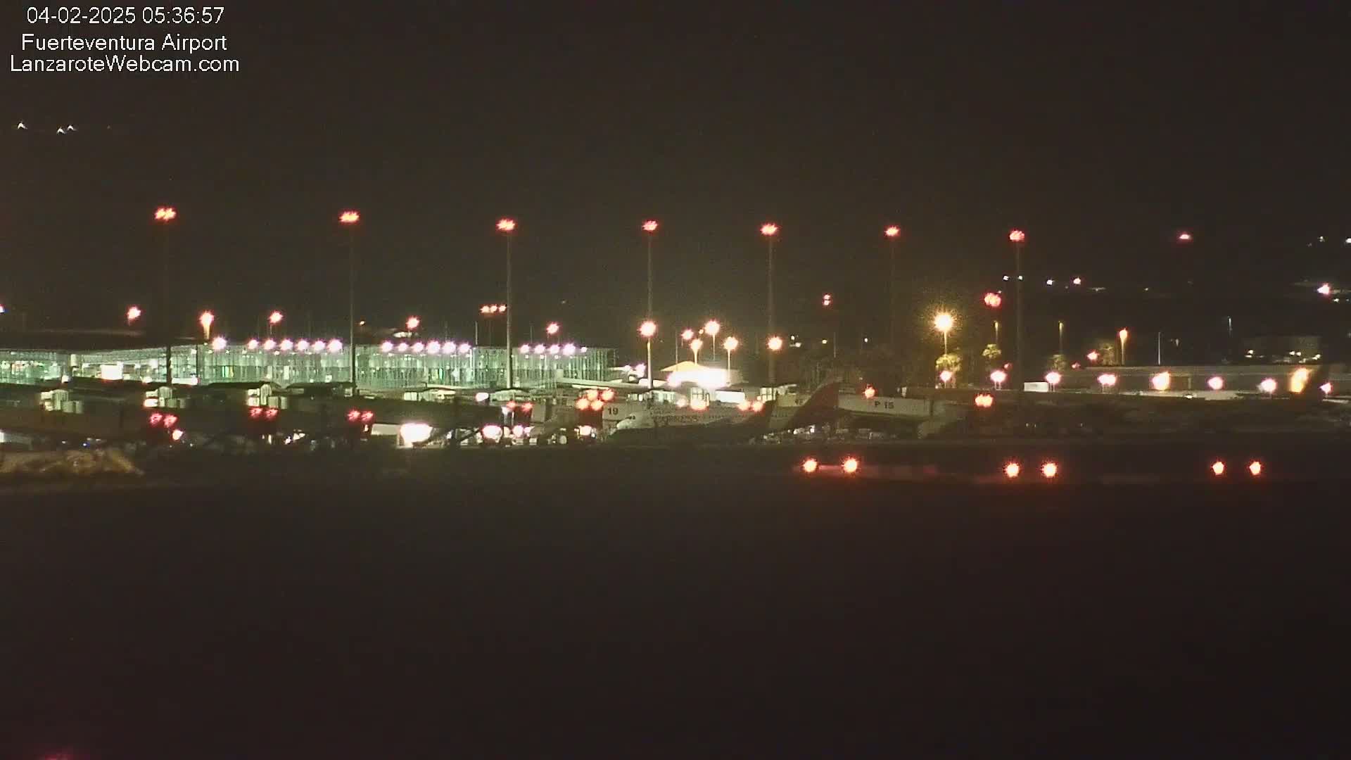 A brightly lit airport at night shows several airplanes parked near a terminal building under a clear, dark sky.