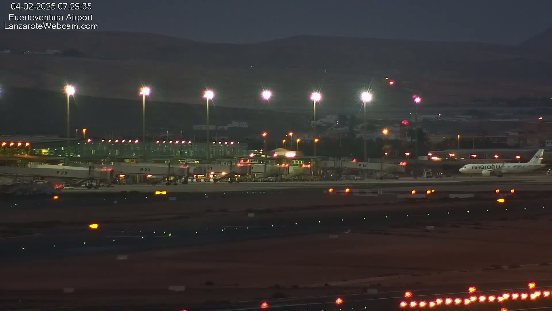 A nighttime view of an airport tarmac showing several planes at gates, with brightly lit jet bridges and tall lights illuminating the scene under a dark sky.