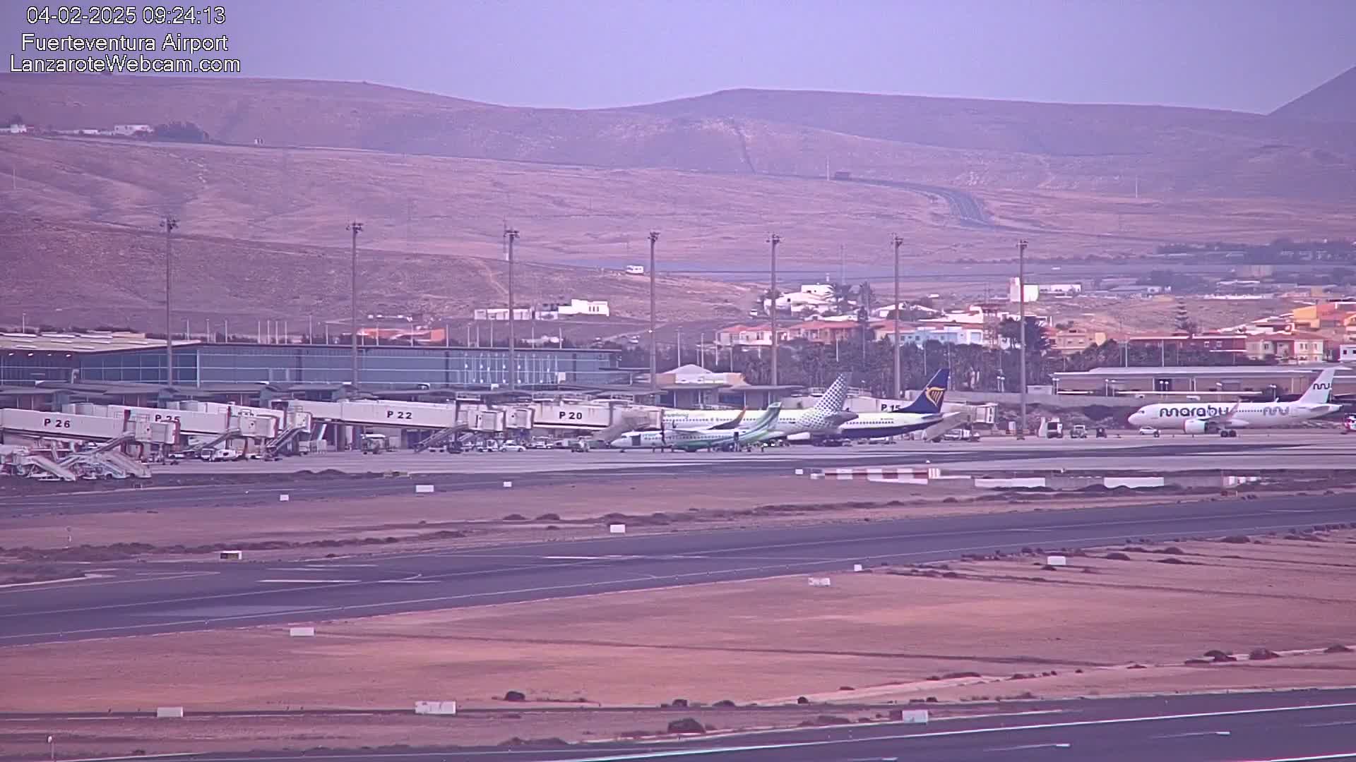 Several airplanes are parked at an airport tarmac with jet bridges connected to some of them, under hazy conditions with mountains in the background.