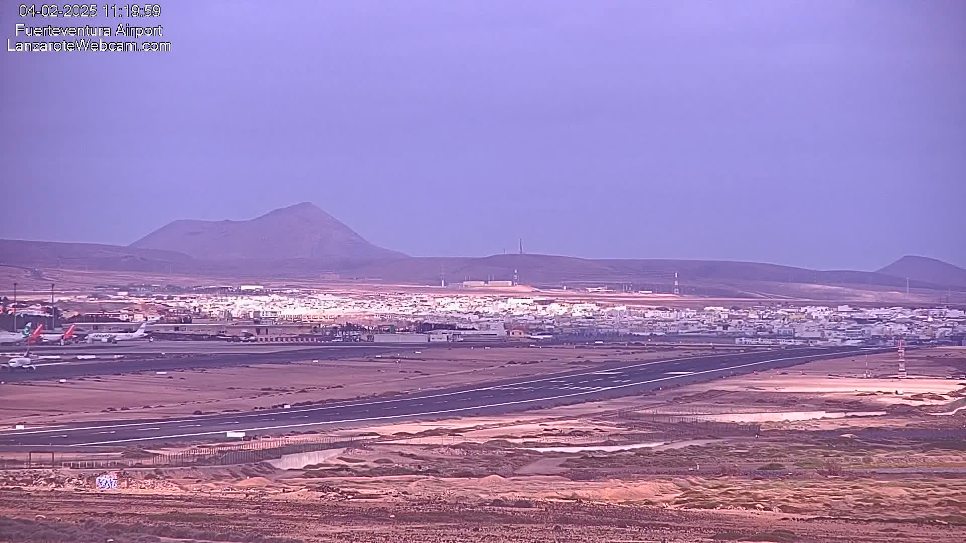 A long runway at an airport is situated in a flat, arid landscape under a hazy purple sky, with low mountains visible in the distance.
