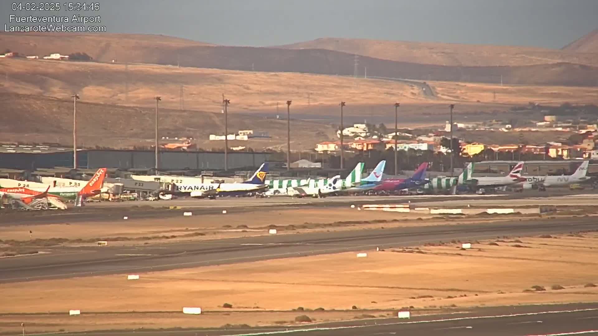 Several airplanes are parked at an airport tarmac under a hazy, sunny sky with a hilly landscape in the background.