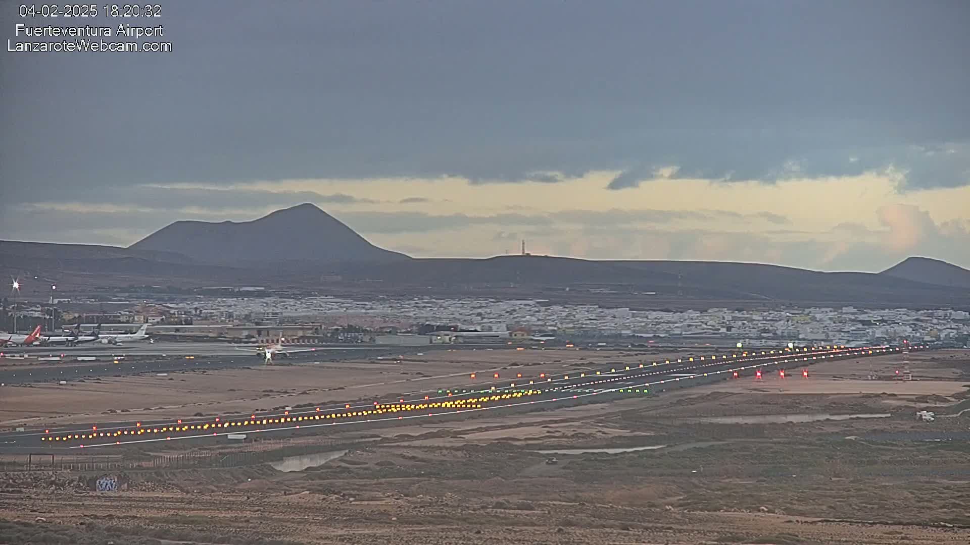 An airport runway with lights illuminated at dusk, under a cloudy sky, with volcanic mountains in the background.
