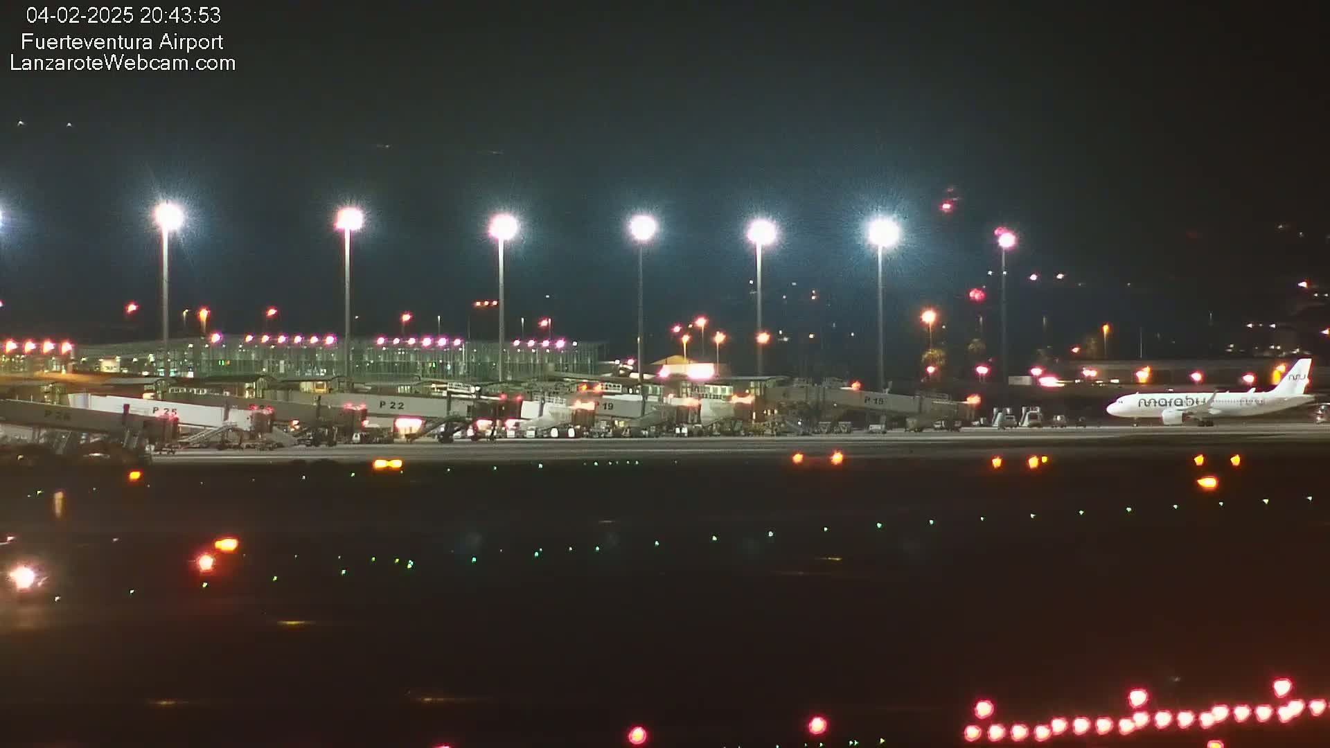 A brightly lit airport at night shows several airplanes near gates and one on the tarmac, under a clear, dark sky.