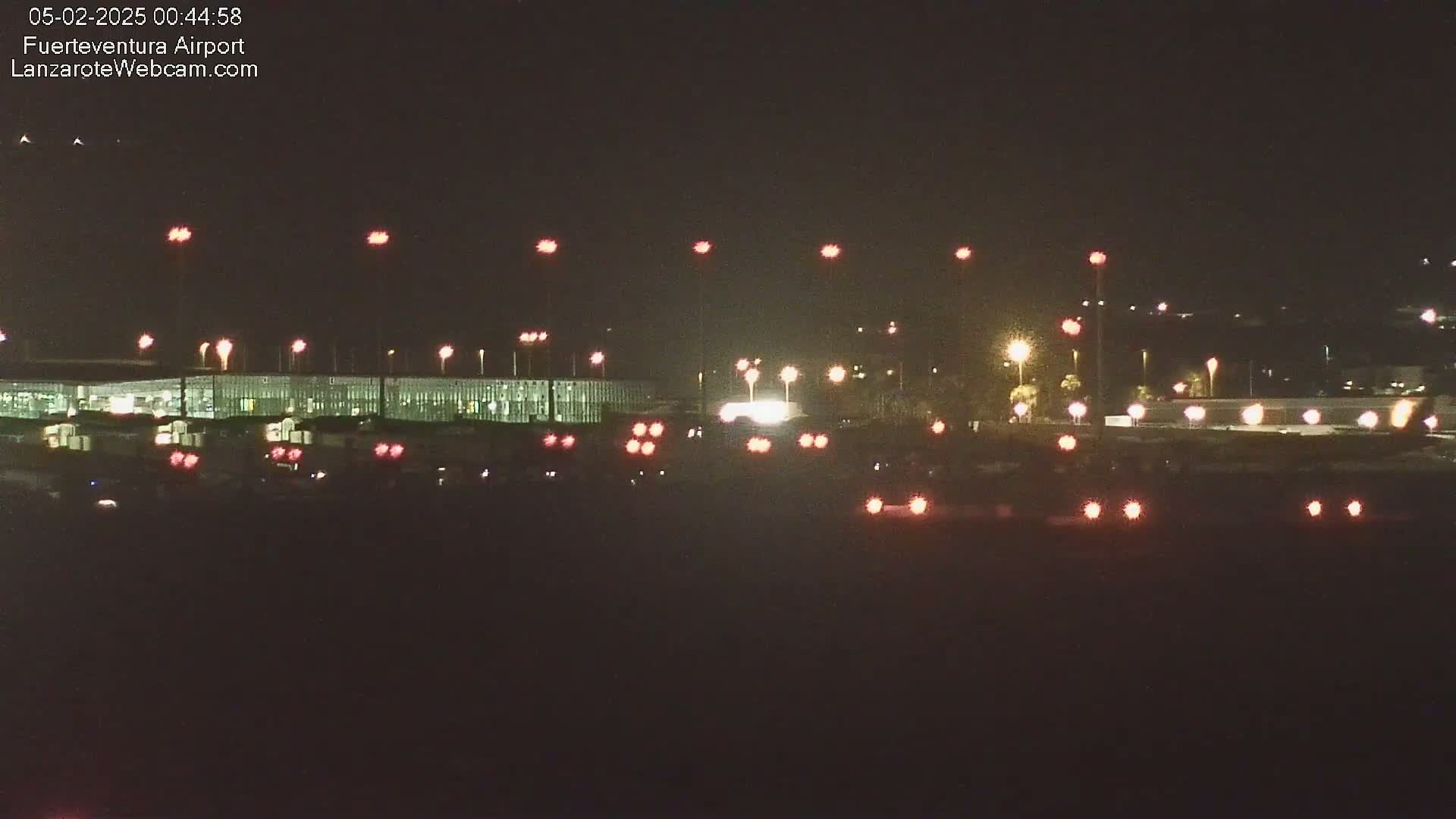 A nighttime view of an airport tarmac with numerous lights illuminating the runway and surrounding buildings.