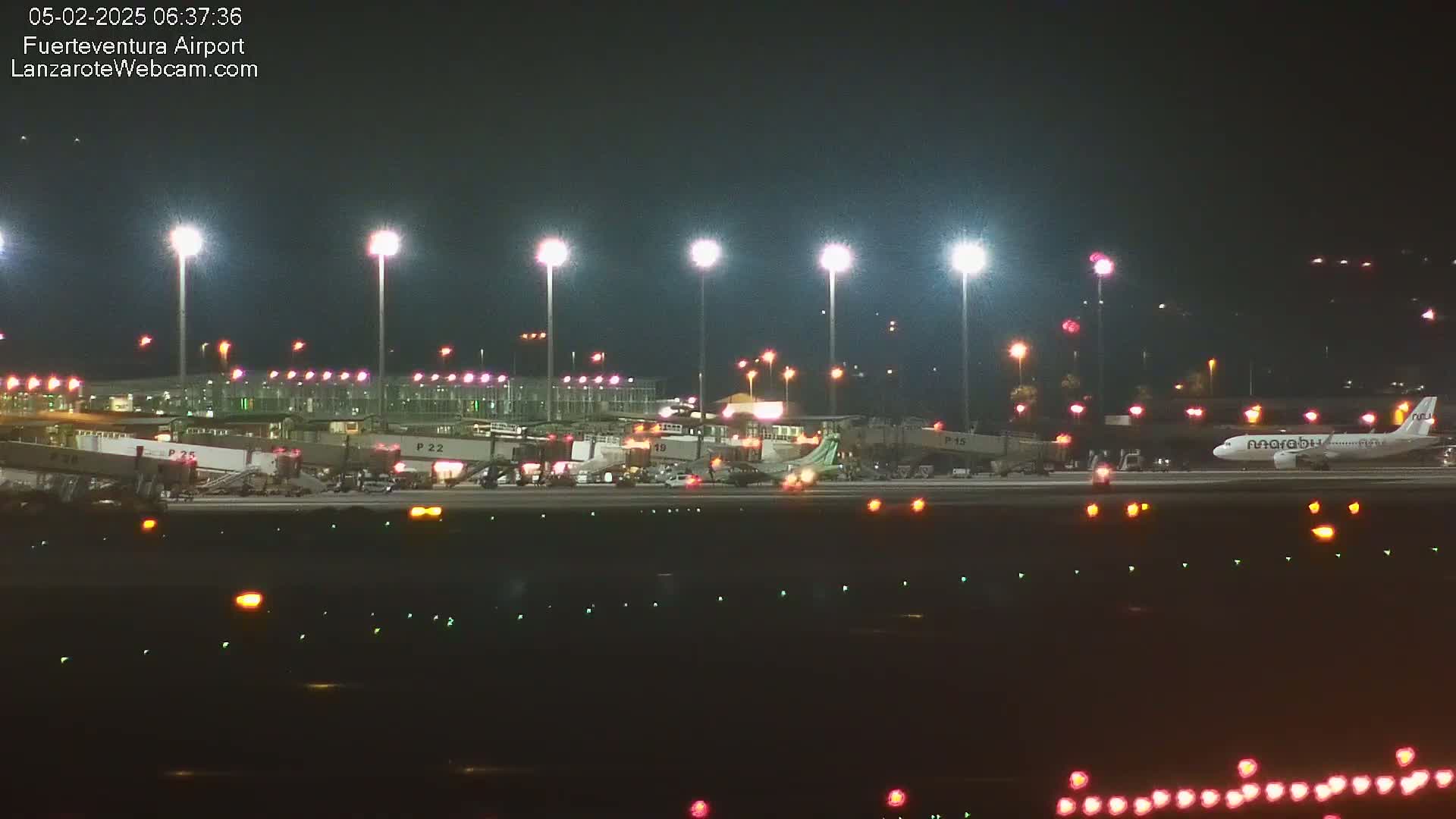 A brightly lit airport tarmac at night shows an airplane parked near several jet bridges and airport buildings.