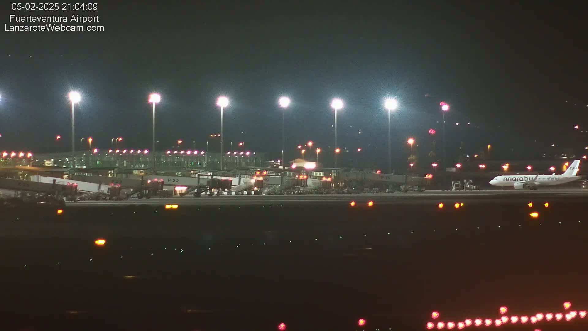 A brightly lit airport at night shows an airplane on the tarmac near several gates and jet bridges.