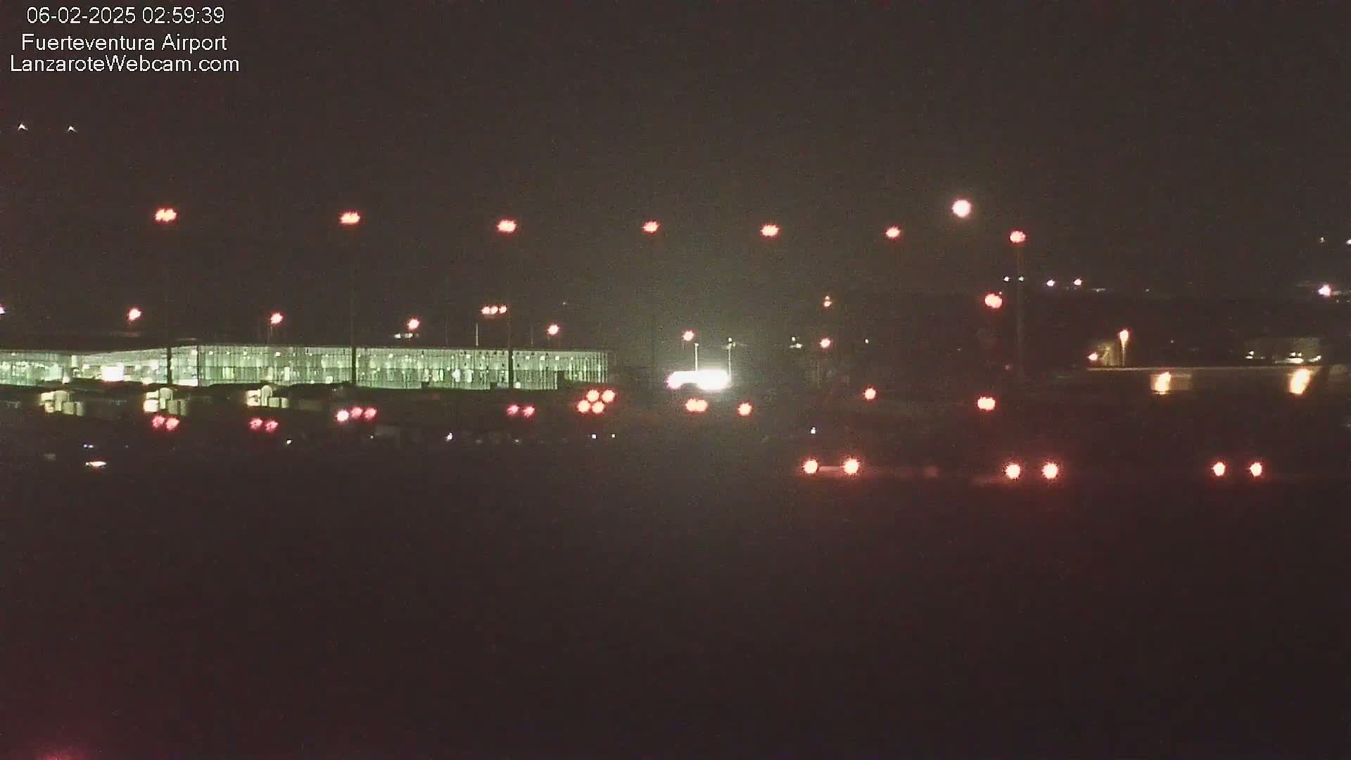 At night, a long, illuminated building is visible in the distance, with numerous red and white lights scattered across the foreground and background.