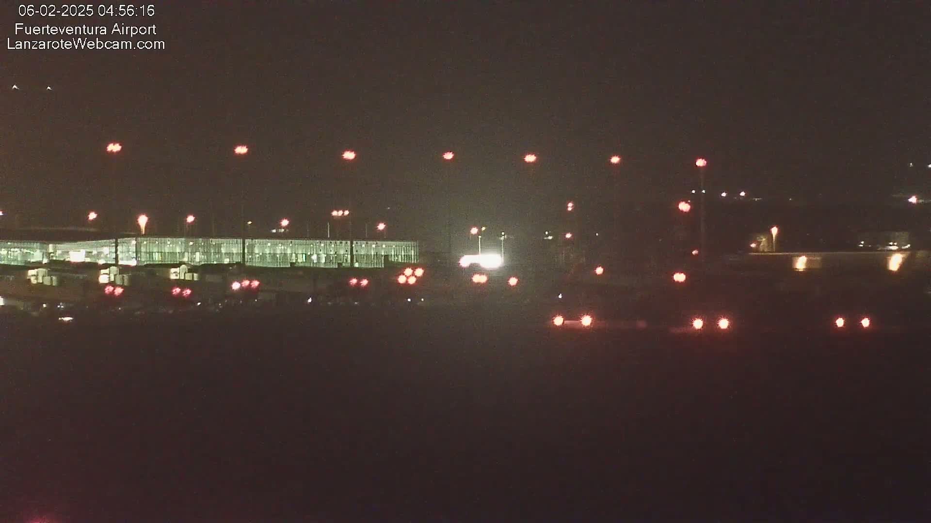 A brightly lit airport terminal at night is visible, with numerous red lights in the foreground and background.