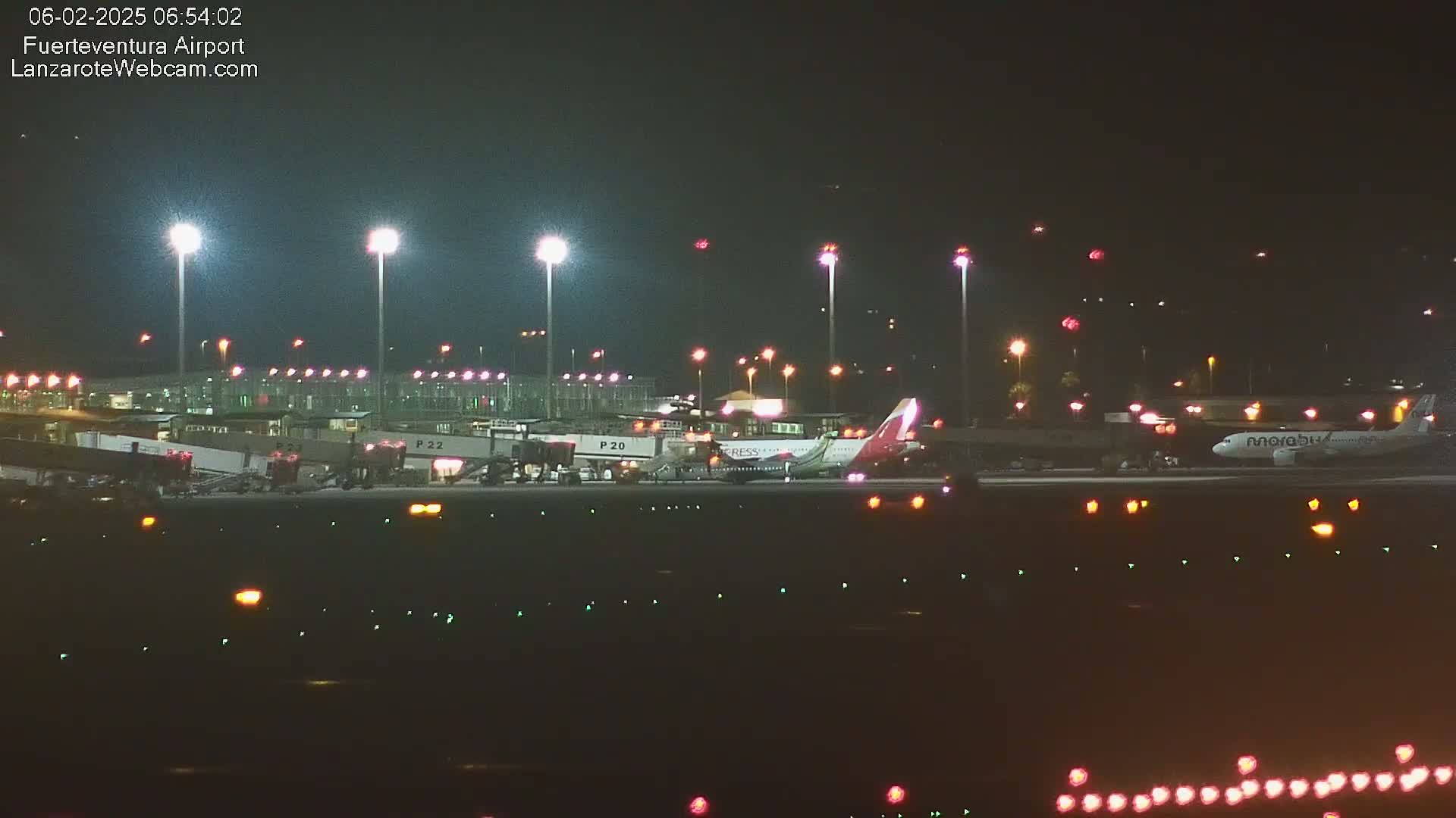 At night, two airplanes are parked at an airport gate surrounded by bright lights and ground support equipment.