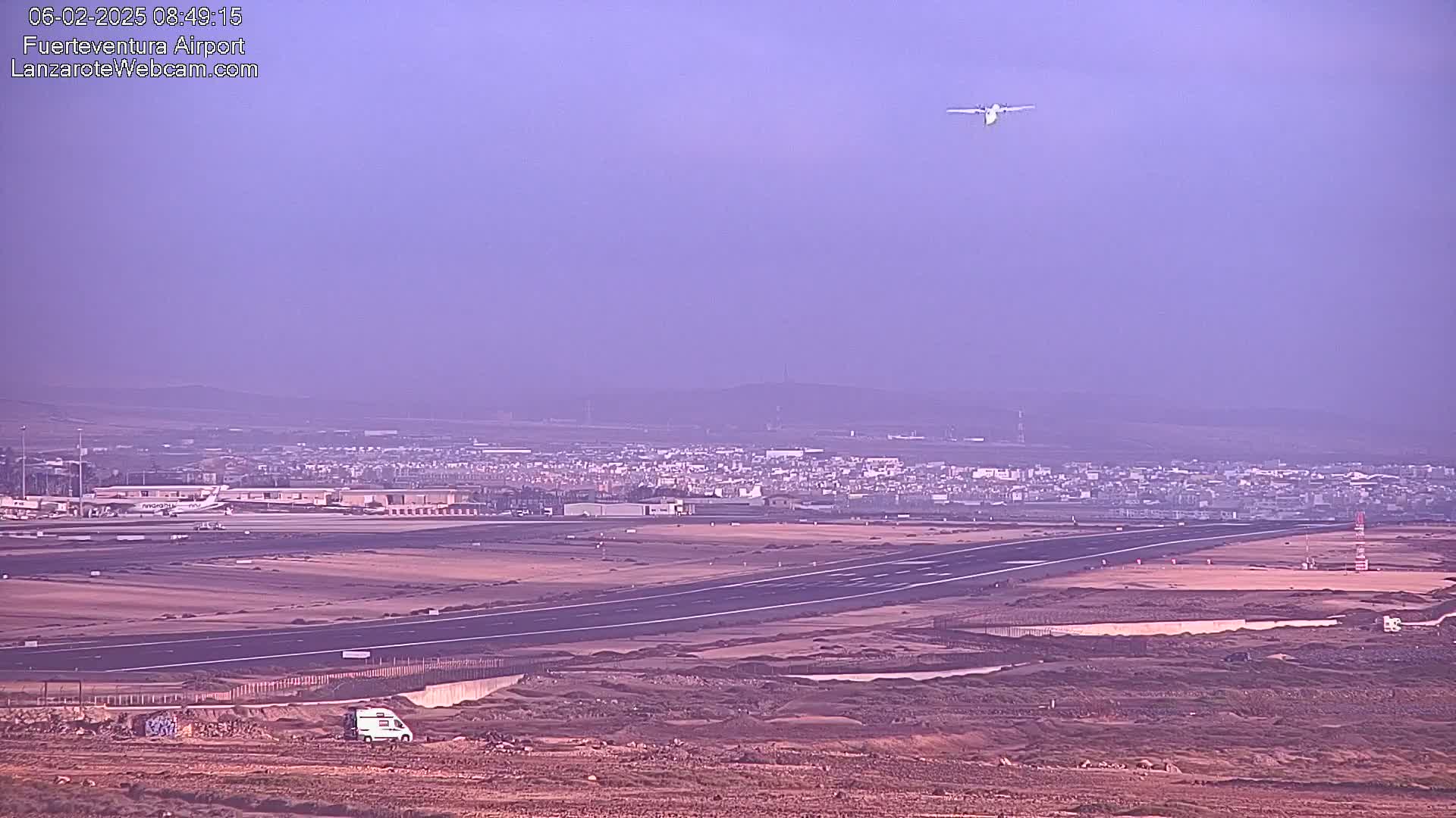 A small airplane flies over an airport runway and surrounding construction areas under a hazy, light purple sky.