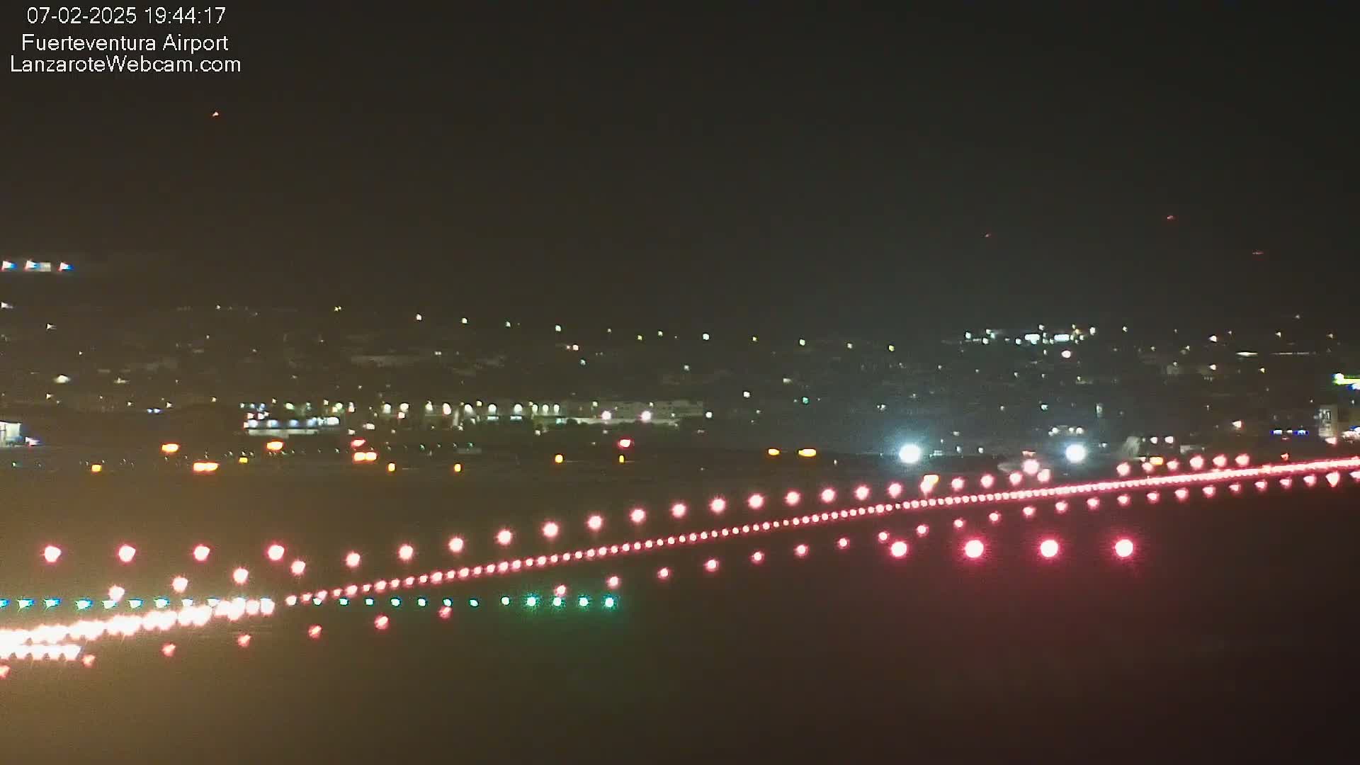A nighttime view of an airport runway with illuminated lights, showing a distant city skyline under a clear, dark sky.
