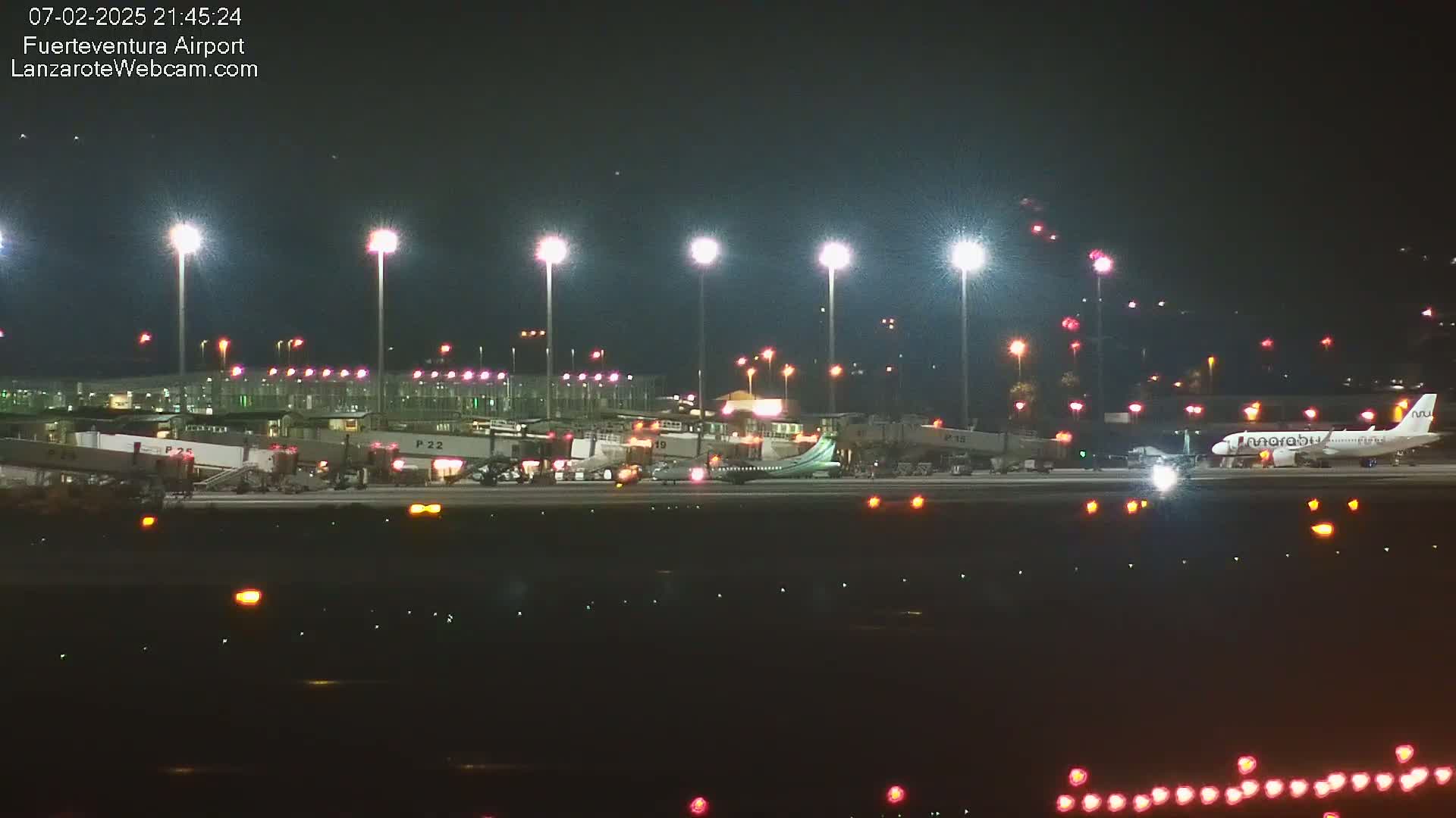 A nighttime view of an airport tarmac with several airplanes, including one at a gate, illuminated by bright lights under a dark sky.