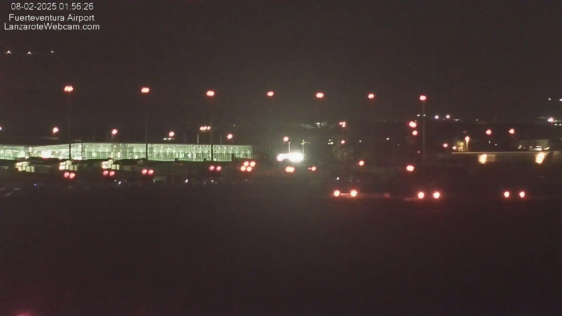 A nighttime long shot of a brightly lit airport terminal and roadway with numerous red taillights visible.
